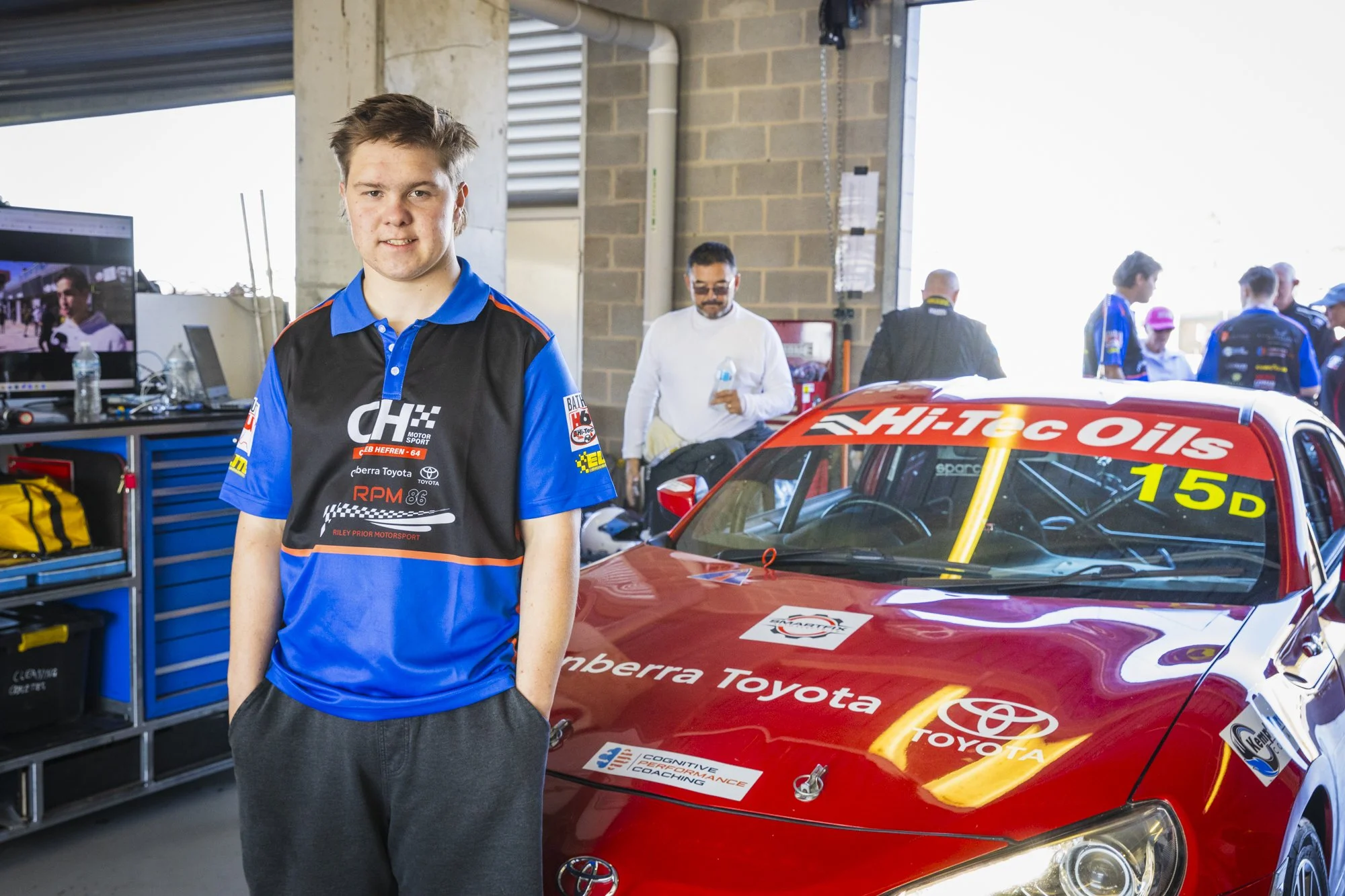 Caleb Hefren - a young man in a racing team uniform standing next to a red race car in a garage or pit area, with people and tools in the background.