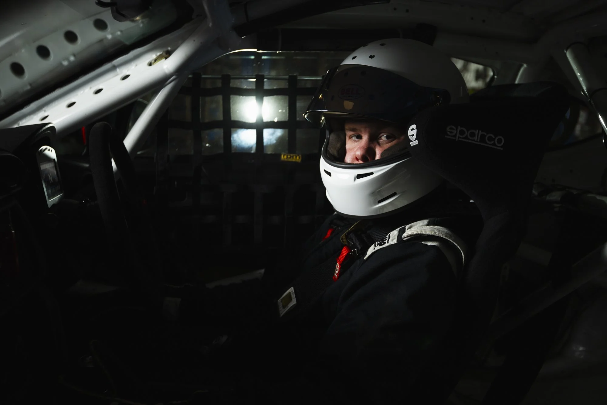 Race car driver wearing a white helmet and racing suit, sitting inside a race car with a black safety cage behind him.