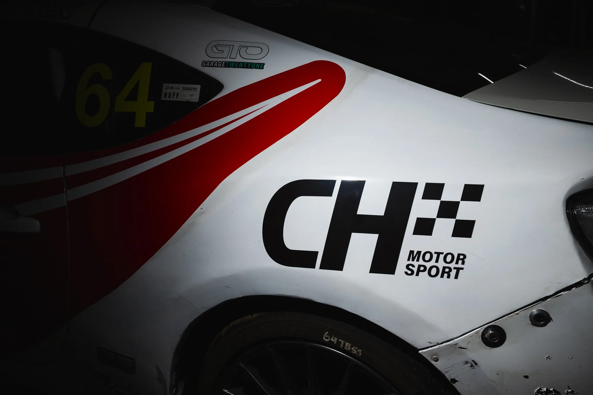 Close-up of a white race car with black and red branding, including the logo 'CH MOTOR SPORT' and the number 64 on the side.