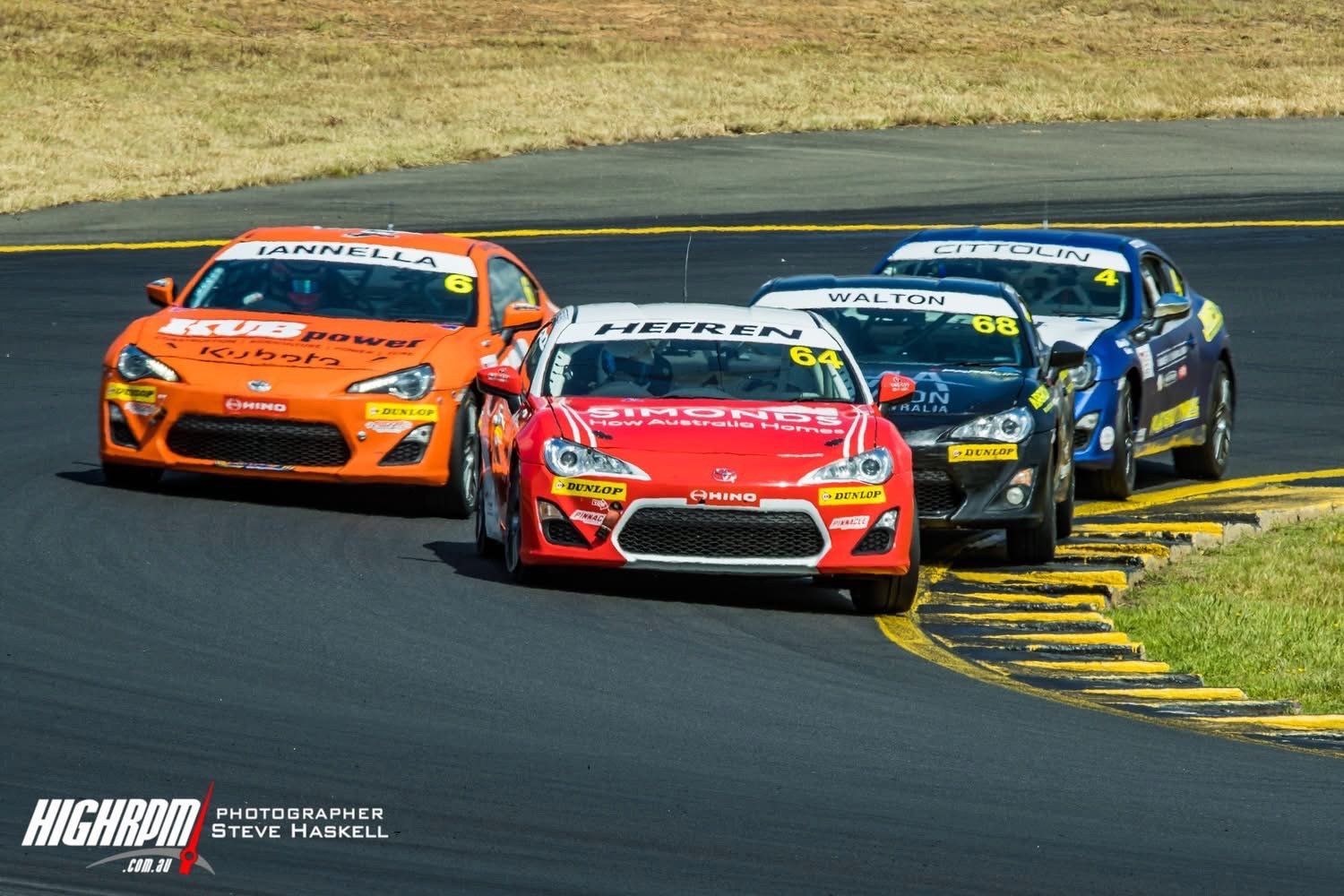 Four race cars navigating a sharp turn on a race track, with safety barriers and grass on the side.