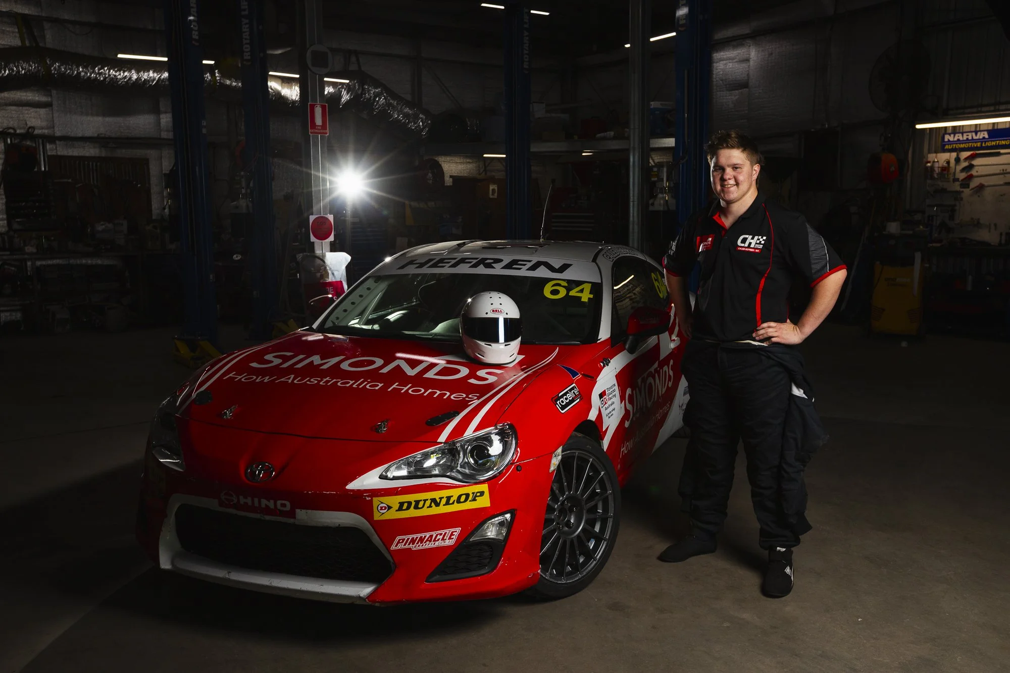 A race car driver in a black racing suit standing next to a red and white race car in a garage.