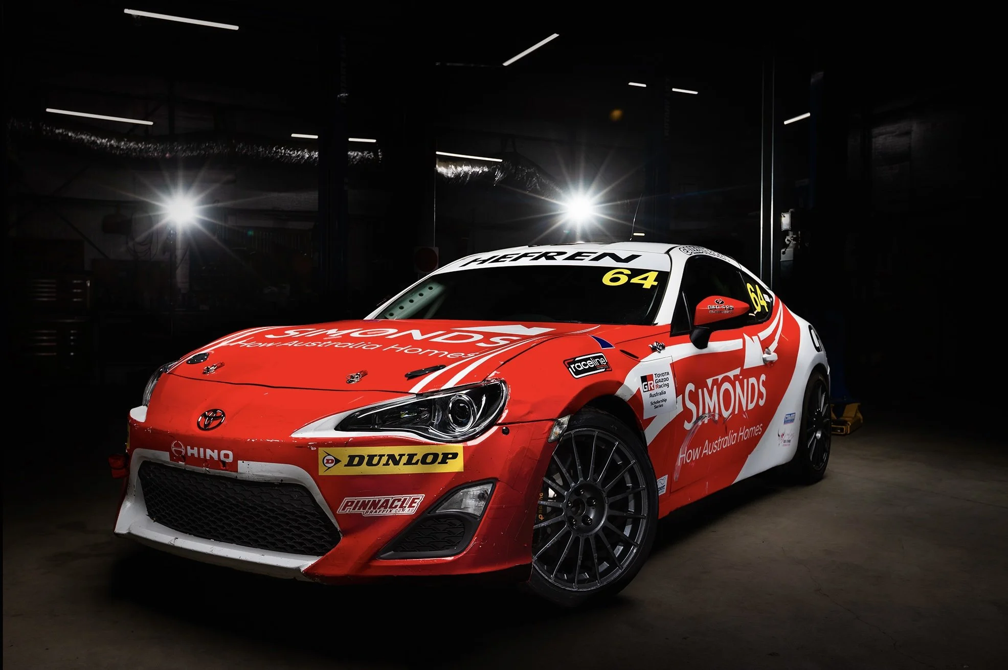 Caleb Hefren's red and white Toyota 86 racing car in a dark garage, illuminated by overhead lights, displaying sponsorship logos and racing decals.