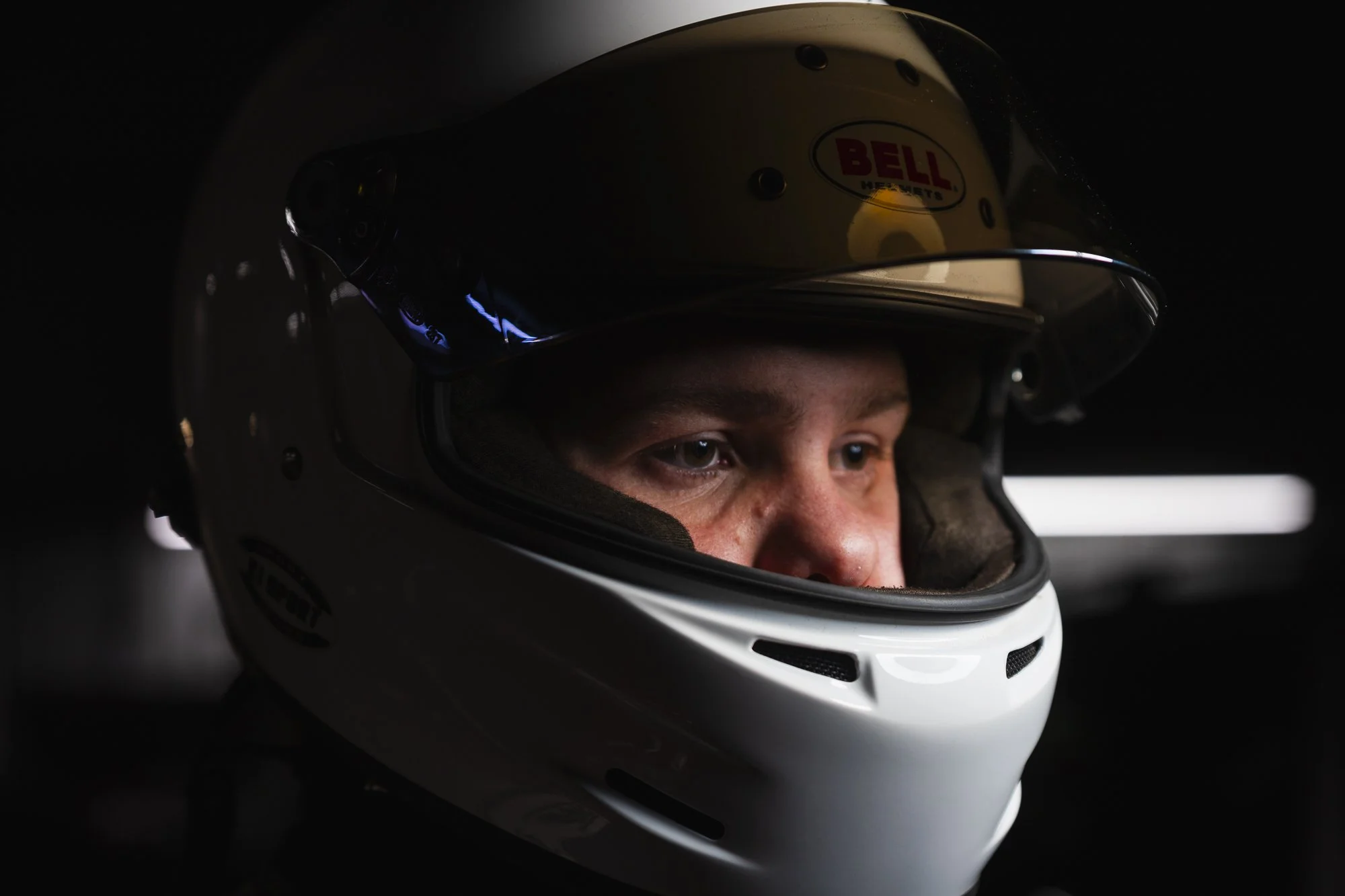 Close-up of a person wearing a white racing helmet with a clear visor, focused expressions, and dark surroundings.