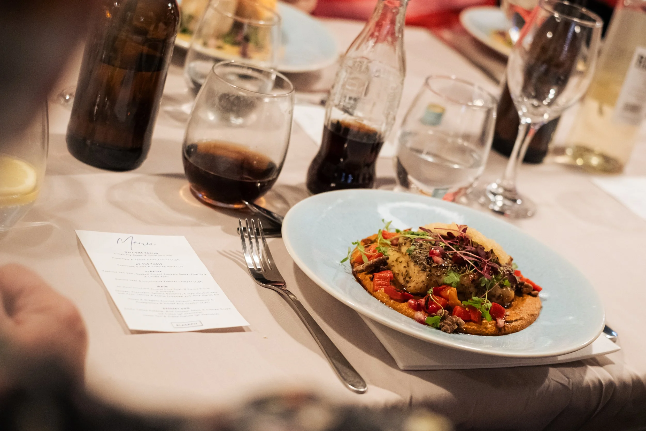 A table setting at a dinner event with a plate of food, drinks, and a menu. The plate contains a gourmet dish garnished with microgreens, served on a blue plate. There are various glasses including wine, water, and soda, along with a bottle of soda a