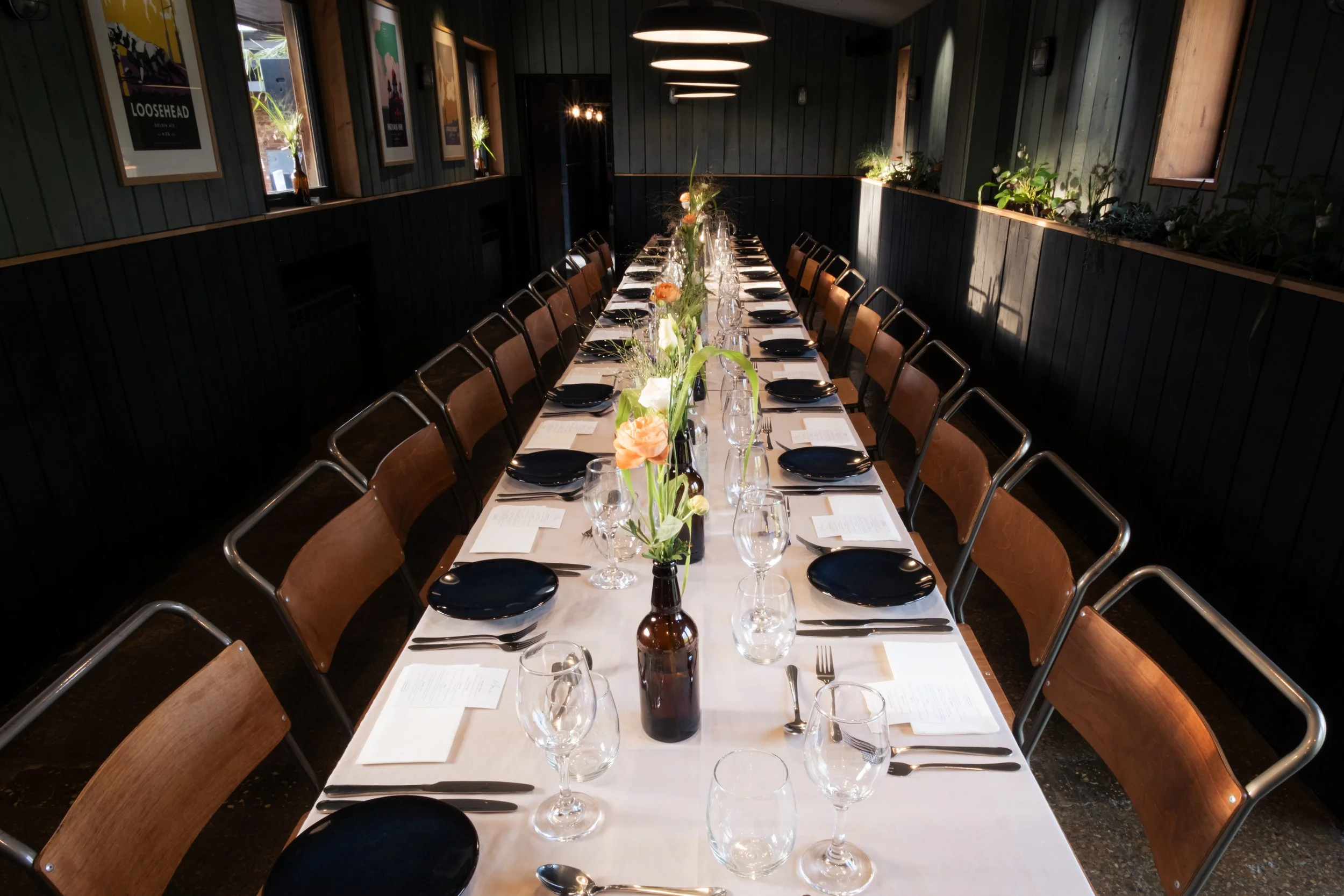 Long dining table set for a formal meal with black plates, wine glasses, silverware, and a white tablecloth, decorated with flowers in bottles in the center.