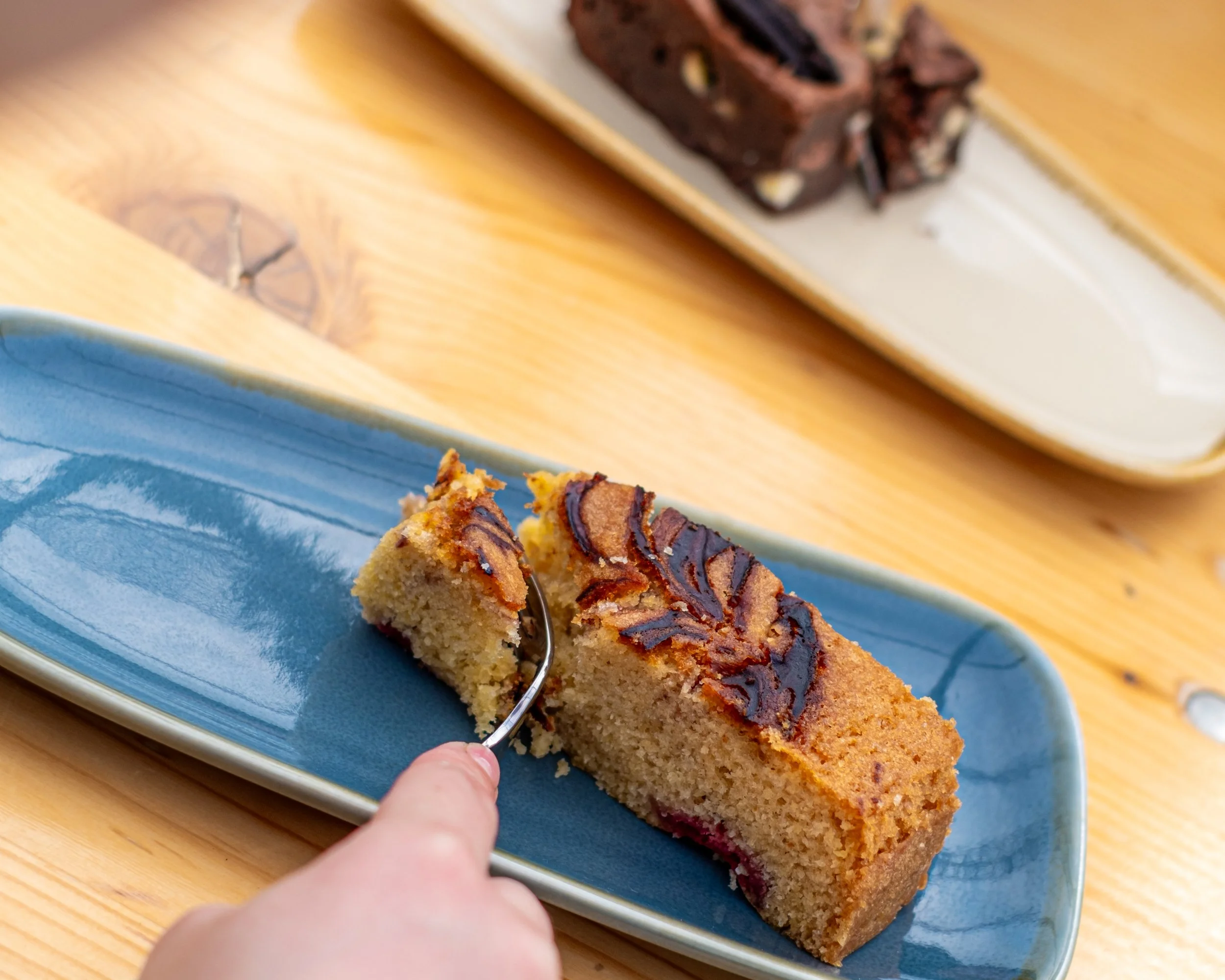 A person is slicing a piece of cake with a fork on a blue platter, with a baked cake with chocolate and nut toppings on a tray in the background Blackpit Café