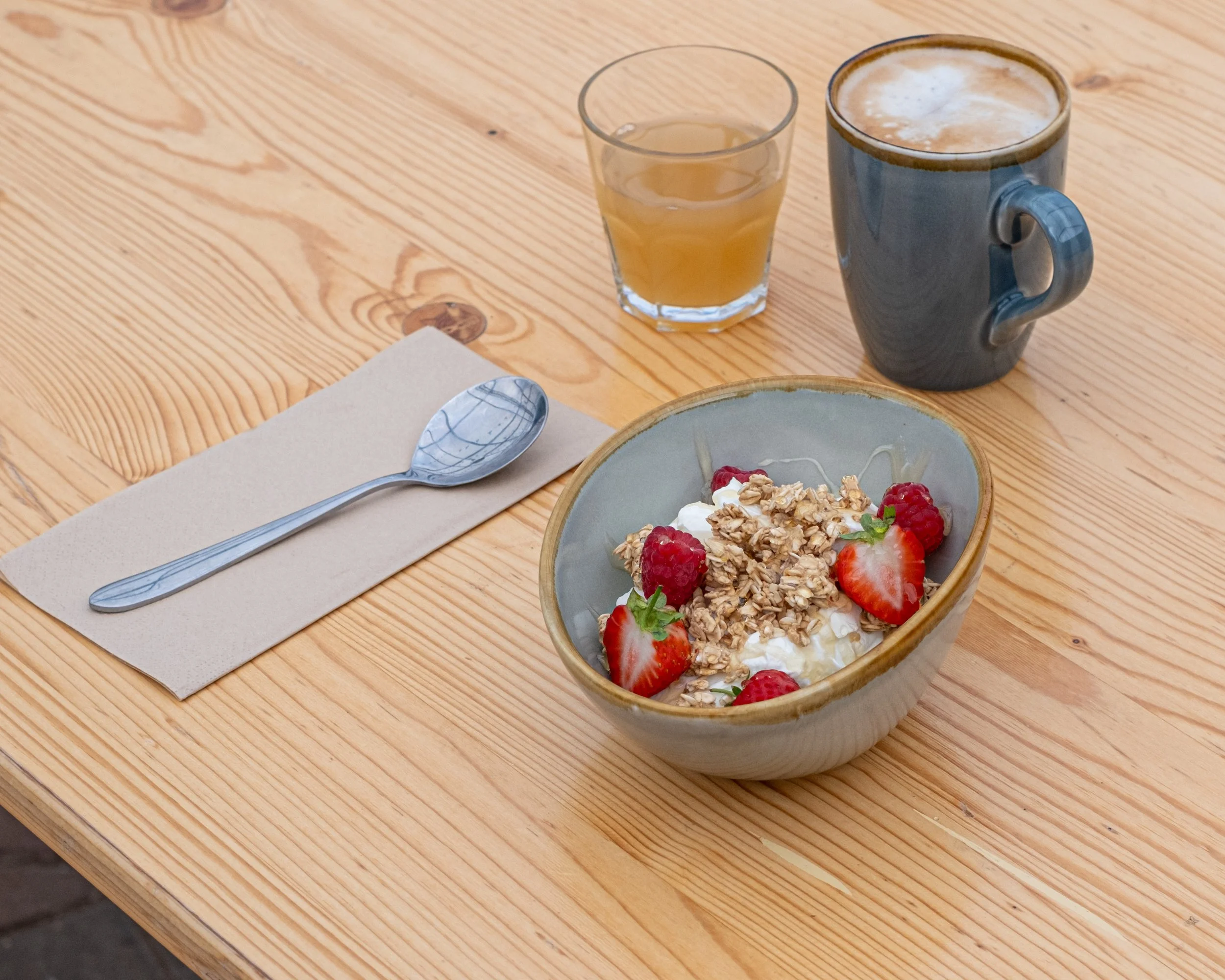 Blackpit Café A breakfast spread on a wooden table including a bowl of yogurt with strawberries and granola, a cup of coffee, a glass of juice, and a spoon on a napkin.