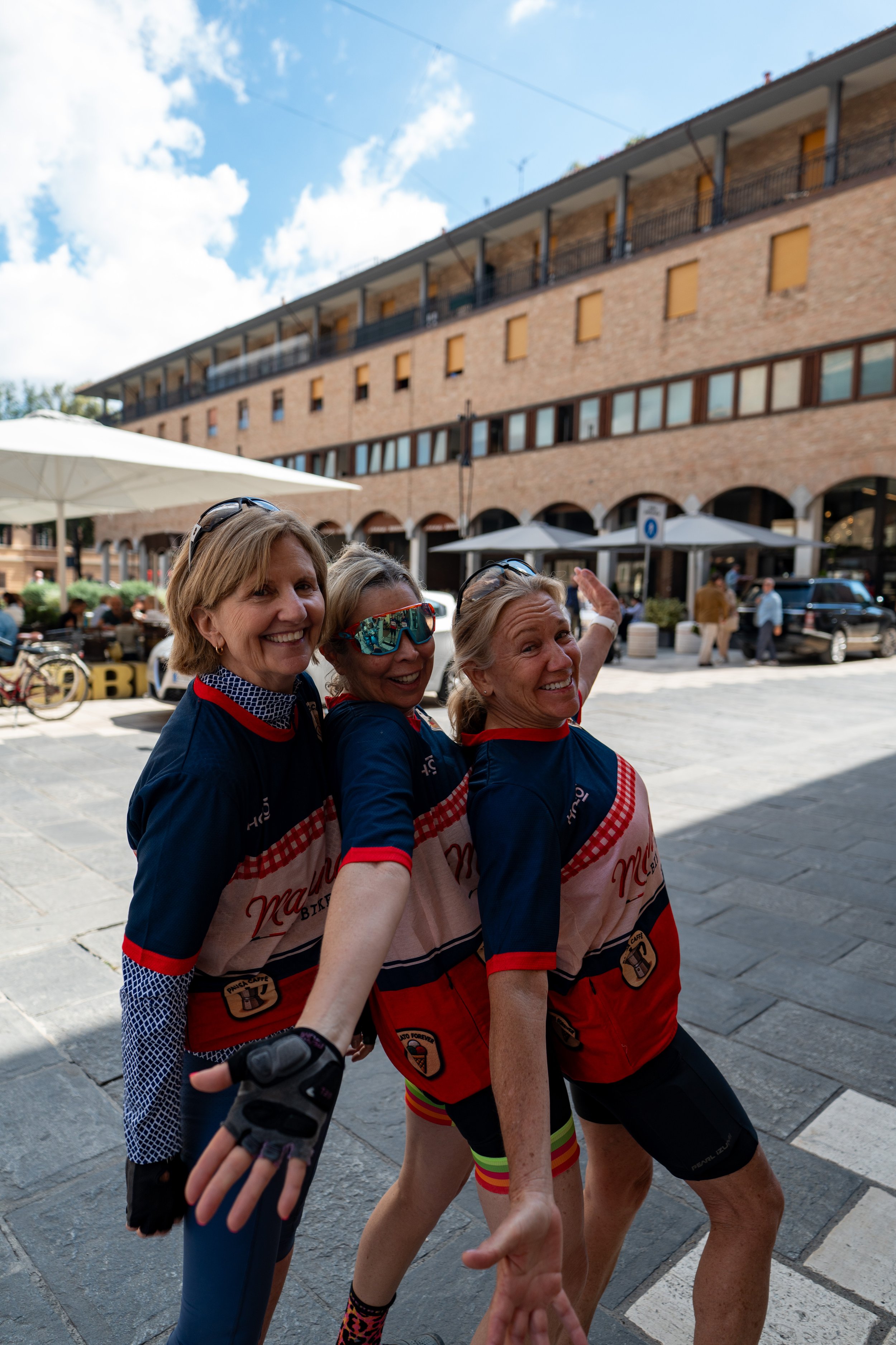 Tre donne sorridenti in abiti sportivi in piedi in una piazza con edifici di mattoni sullo sfondo, durante una giornata di sole.