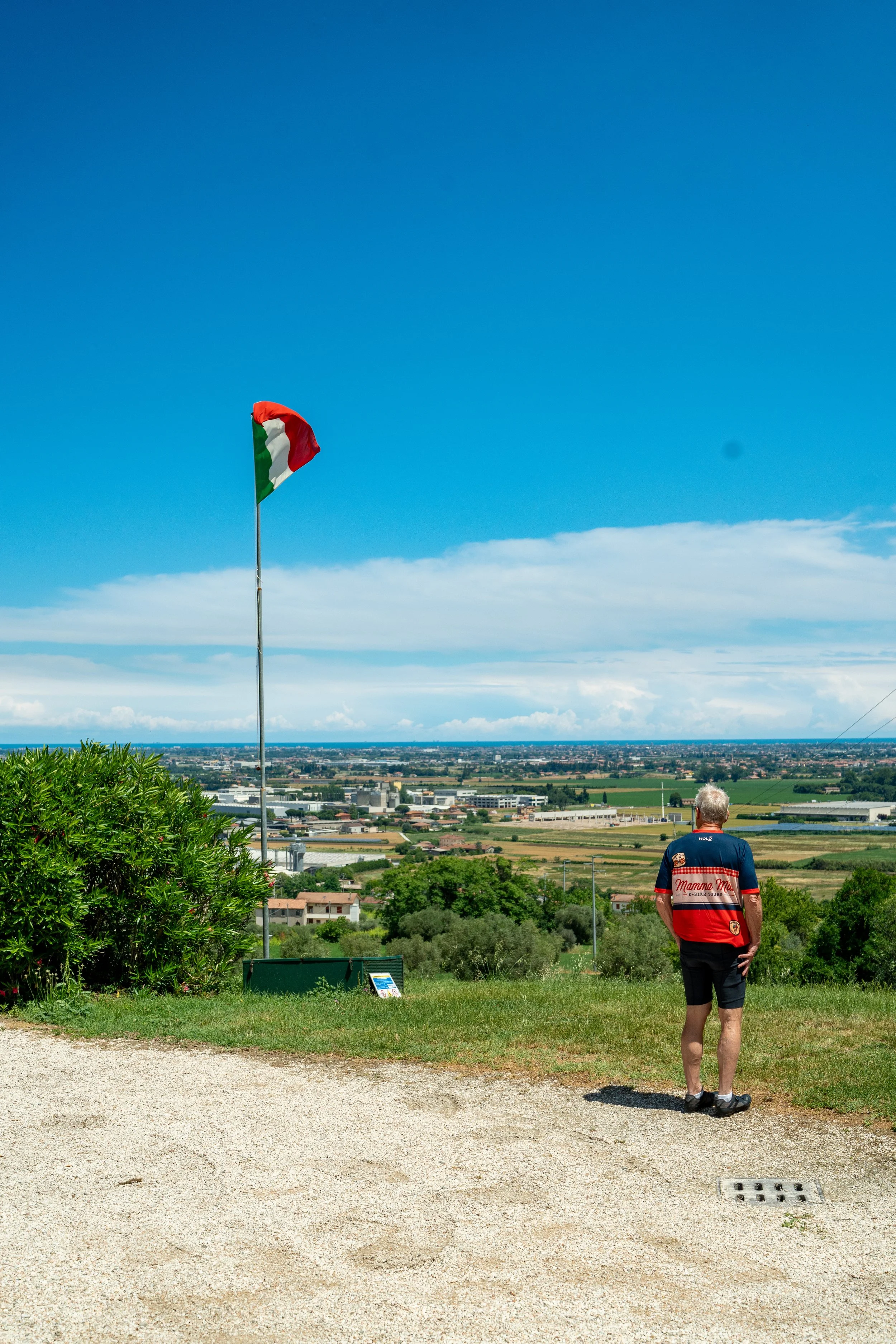 Uomo vestito con maglietta e pantaloncini cammina lungo un sentiero rivolgendo le spalle alla fotocamera, con un paesaggio rurale e un cielo blu sullo sfondo. Sul lato sinistro, un albero verde, e a sinistra un'asta con una bandiera italiana svolazzante.