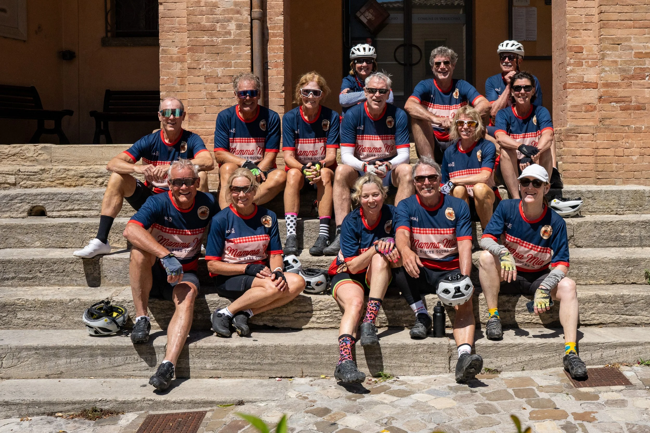 Gruppo di persone in abbigliamento sportivo sedute e in piedi su una scalinata di pietra davanti a un edificio in mattoni, indossando caschi, occhiali da sole e guanti, pronti per una gita in bicicletta.