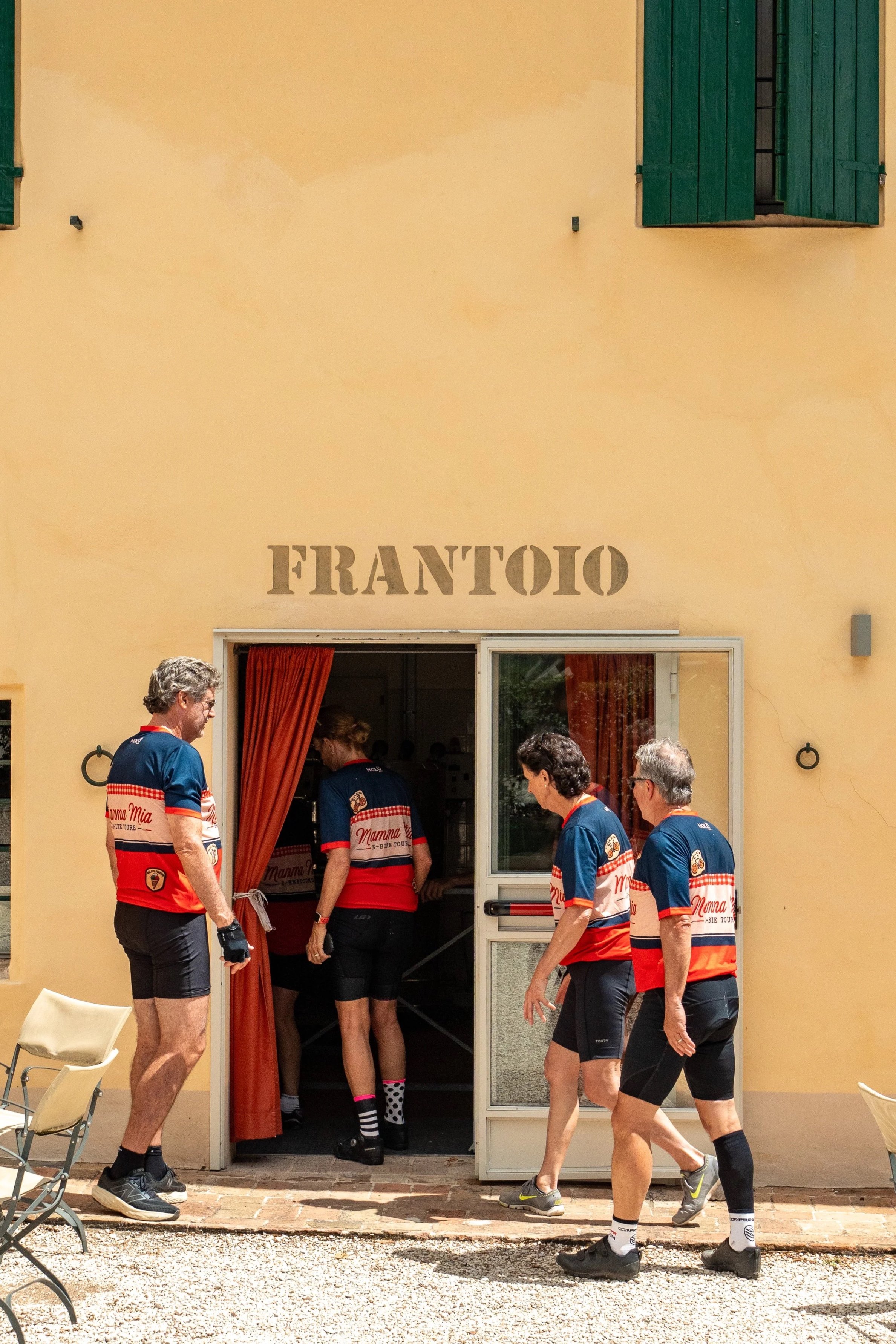 Gruppo di persone in abbigliamento sportivo che entra in un locale con la scritta 'FRANTOIO' sopra l'ingresso, sulla facciata di un edificio con pareti gialle e finestre con persiane verdi