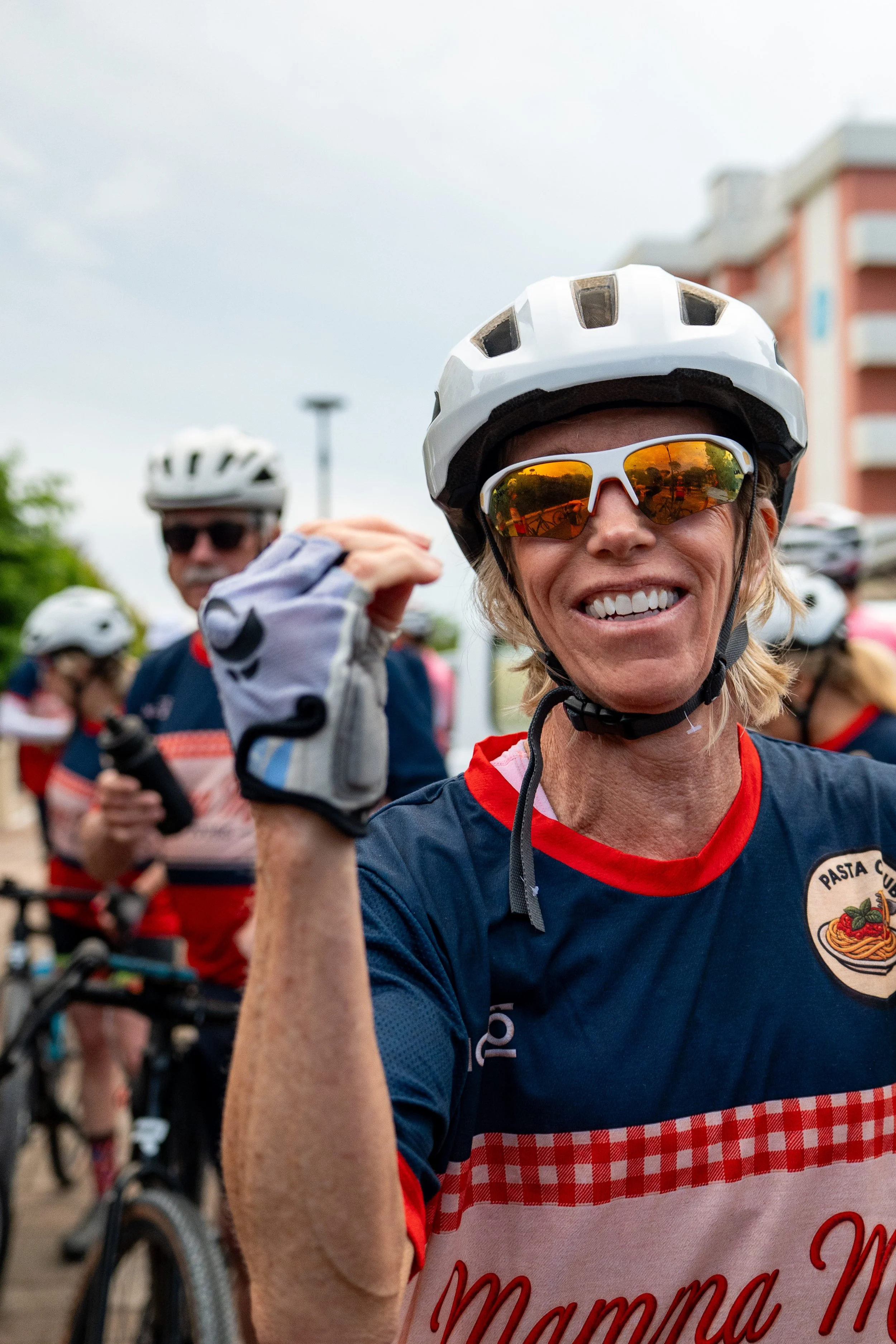 Donna sorridente con casco e occhiali da sole, parte di un gruppo di ciclisti, che indossa una maglietta con la scritta 'Mamma M'.