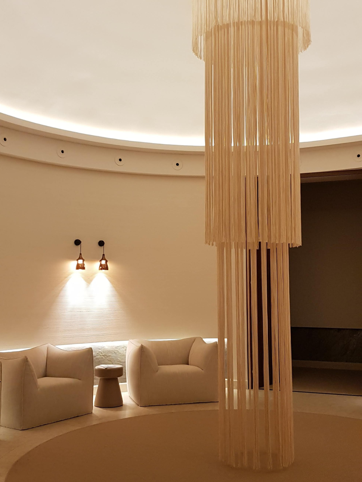 Elegant lobby with beige armchairs and a small side table, illuminated wall lights, and a large wooden cylindrical sculpture or chandelier.