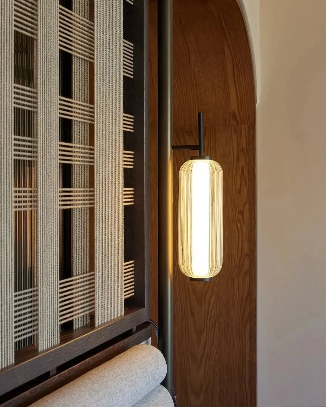Close-up of a modern wall-mounted light fixture with a cylindrical, ribbed glass shade glowing, set against a wooden wall and adjacent decorative lattice panel.