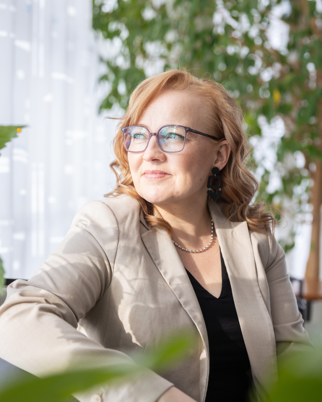 Nainen katseli ikkunasta, selkeä profiili, harmaassa puvussa, lasit ja koruja, sisätiloissa luonnonvalossa, vihreitä kasveja taustalla.