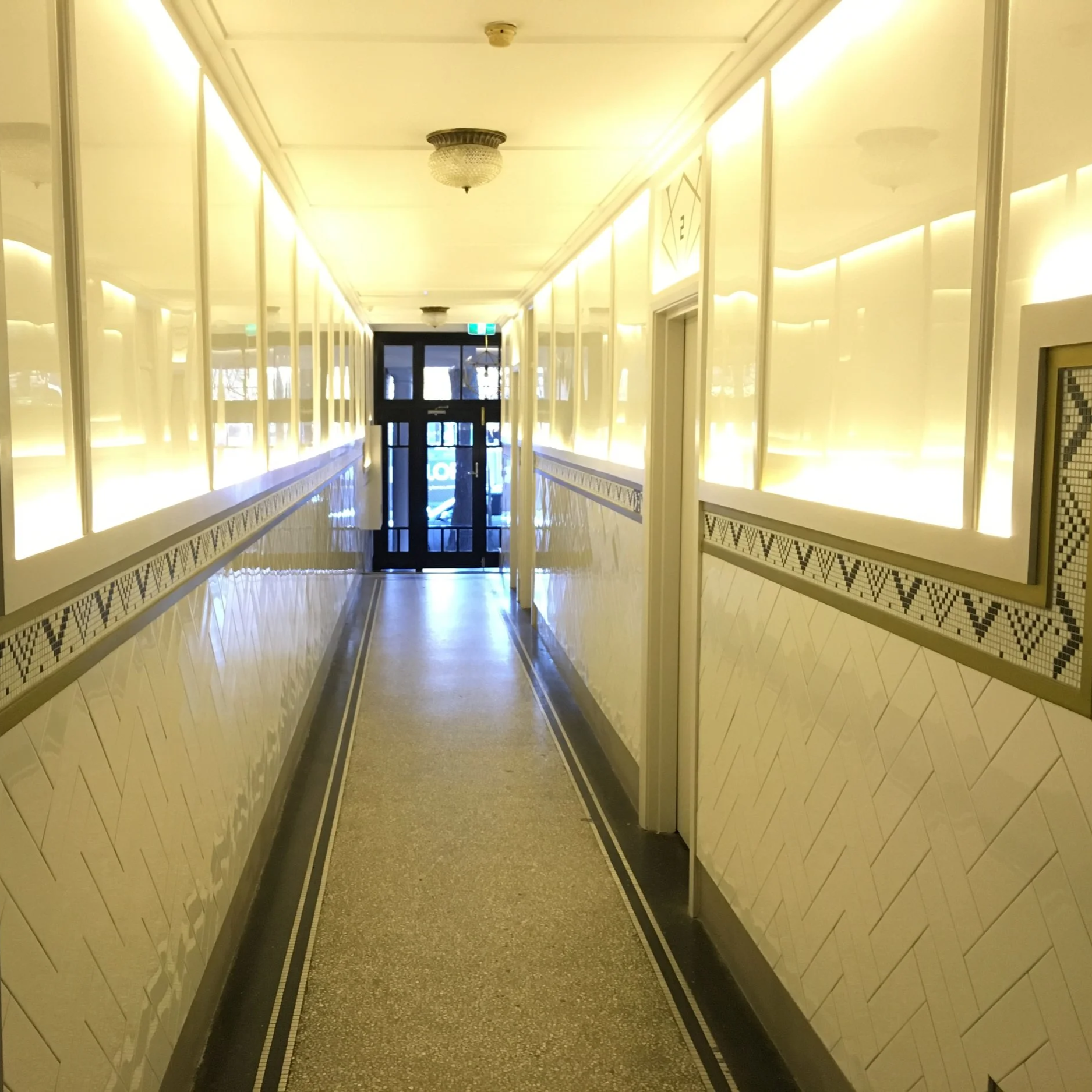 Empty hallway with tiled walls, lit by warm yellow lighting, leading to a glass door at the end.