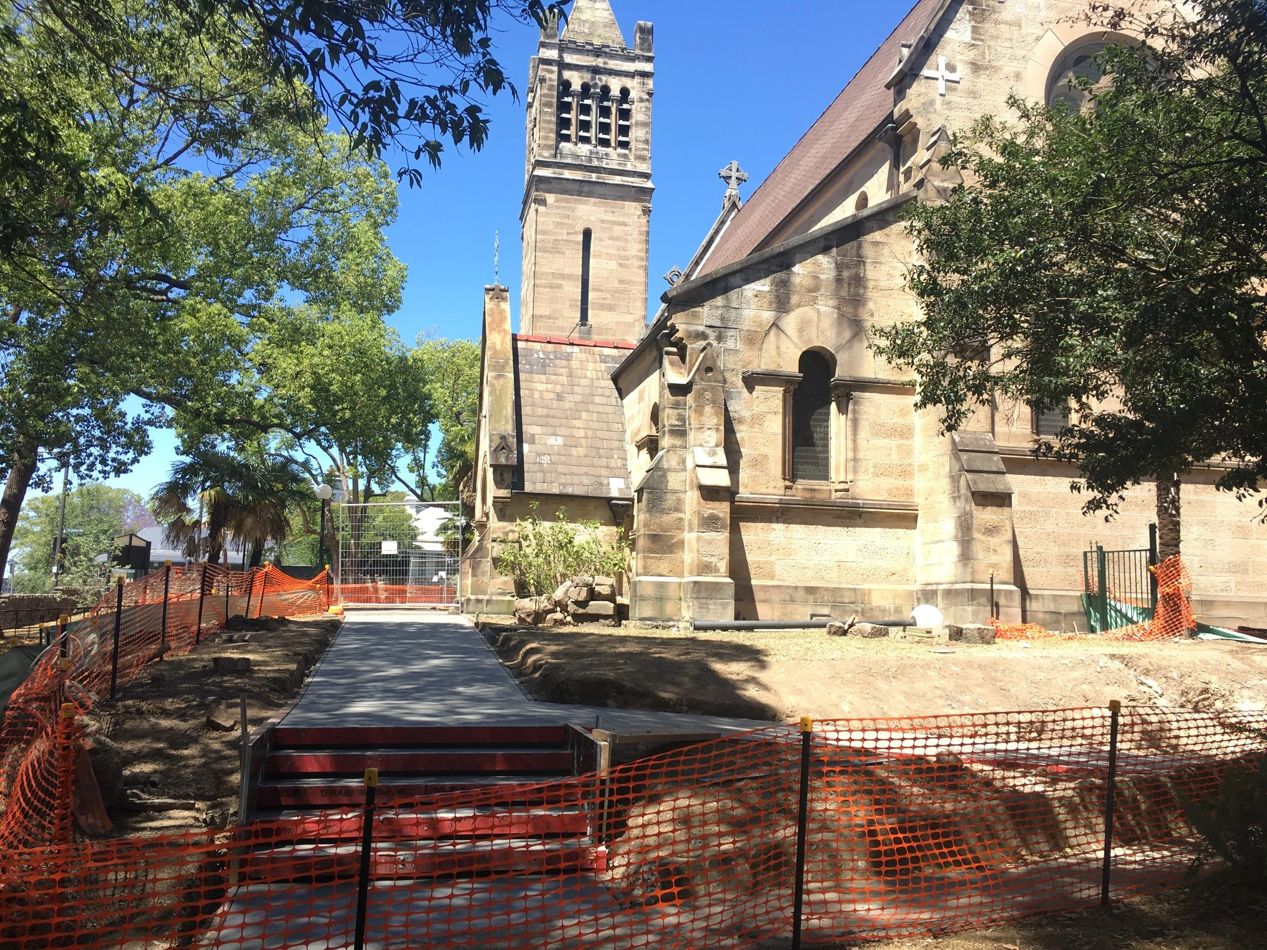 Construction site with a newly paved pathway leading to a stone church building, orange safety fencing, trees, and blue sky.