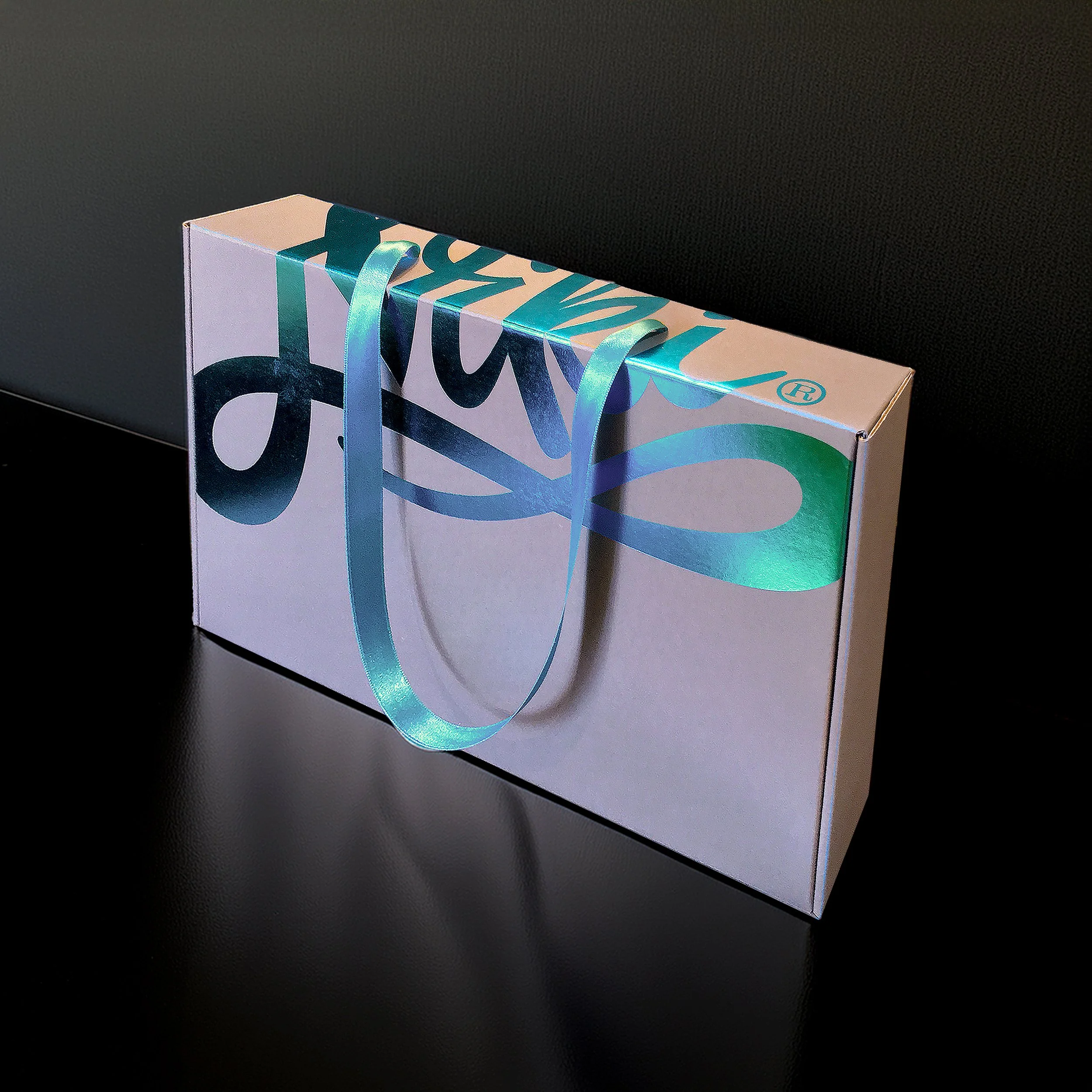 A white rectangular gift box with blue script writing and iridescent ribbon handles, placed on a black surface with a dark background.