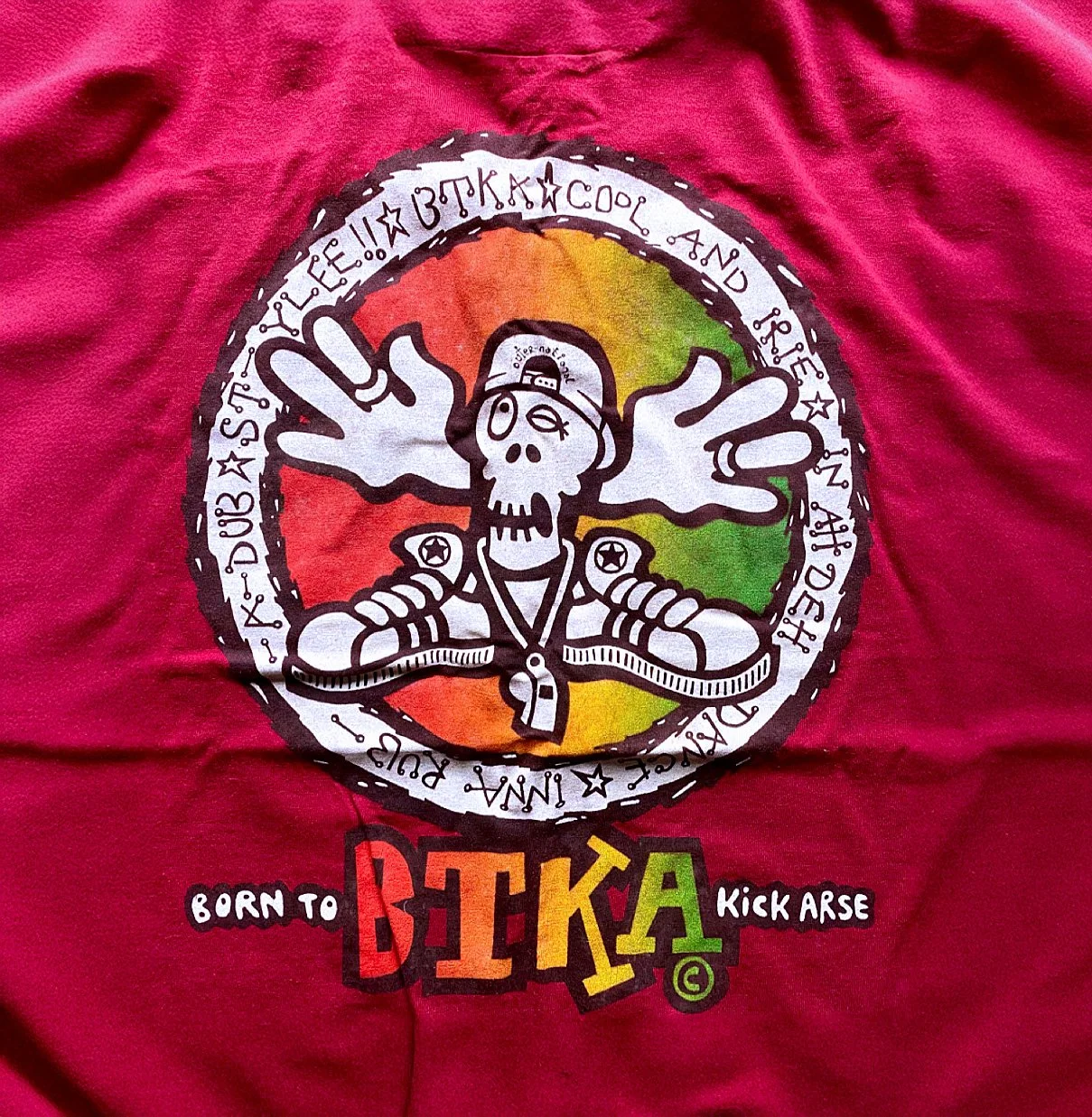 A red fabric with a colorful graphic design featuring a cartoon skeleton with outstretched hands, wearing headphones, inside a multicolored circle with text around it. Below the graphic, the words "Born to B-T-K-A Kick Arse" are printed in colorful letters.