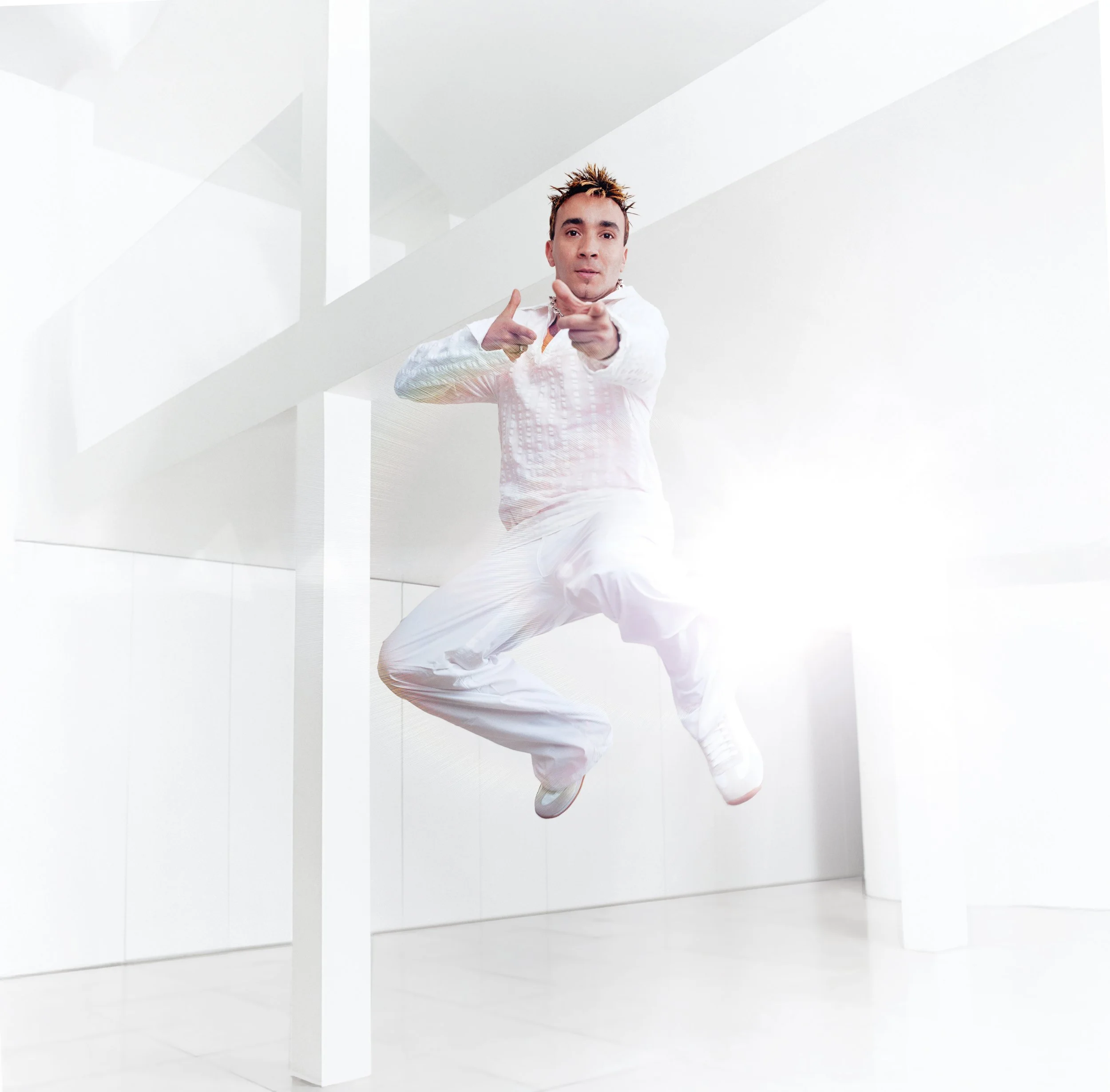 A person with spiked hair dressed in white jumpsuit is jumping in a modern white interior, pointing towards the camera.