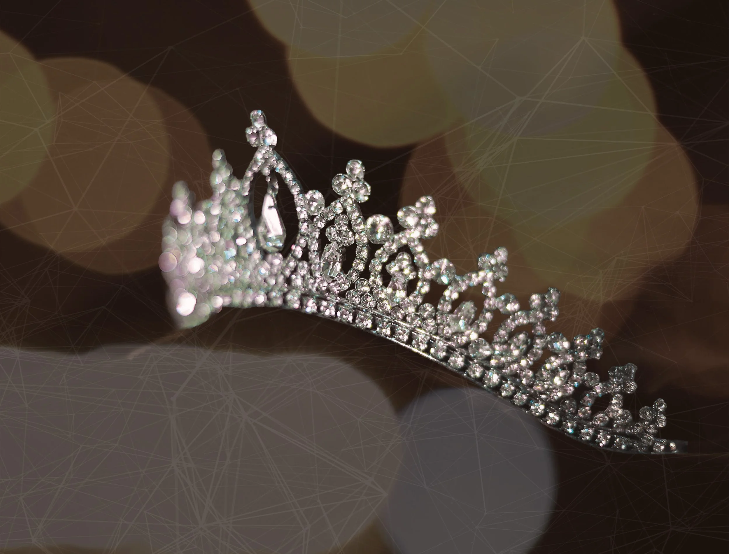 A close-up photo of a silver tiara decorated with small, sparkling rhinestones, placed on a dark, blurred background with circular light bokeh effects.