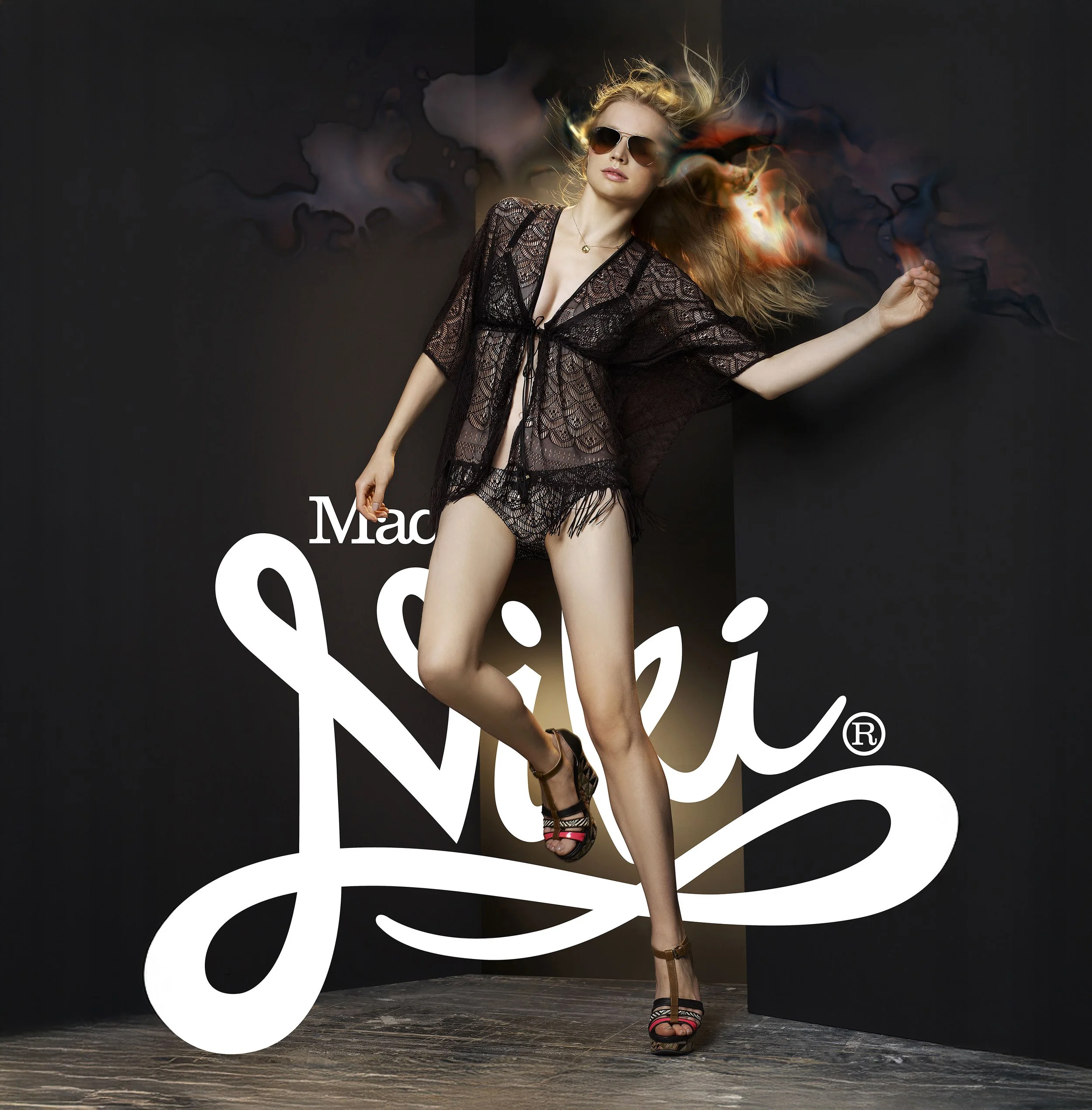 A woman is posing in front of a black background with a large white logo, wearing sunglasses, a black lace cover-up, and high-heeled sandals, with artistic flames coming from her head.