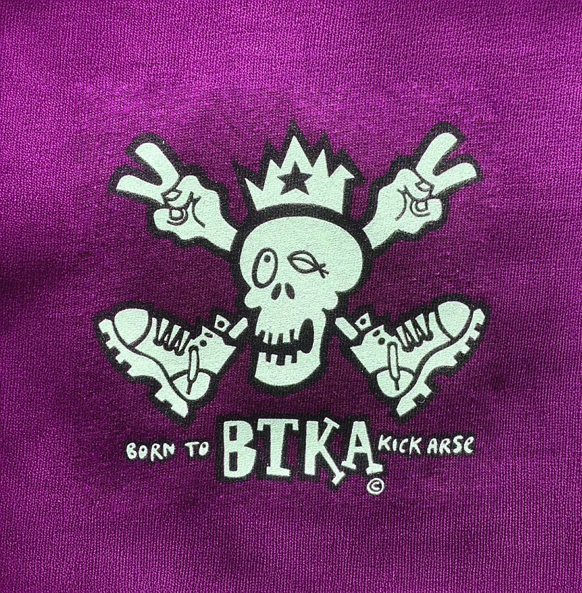Graphic featuring a skull wearing a crown with crossed skateboards, surrounded by sneakers, on a purple background. Text reads "Born to BIKA Kick Arse."