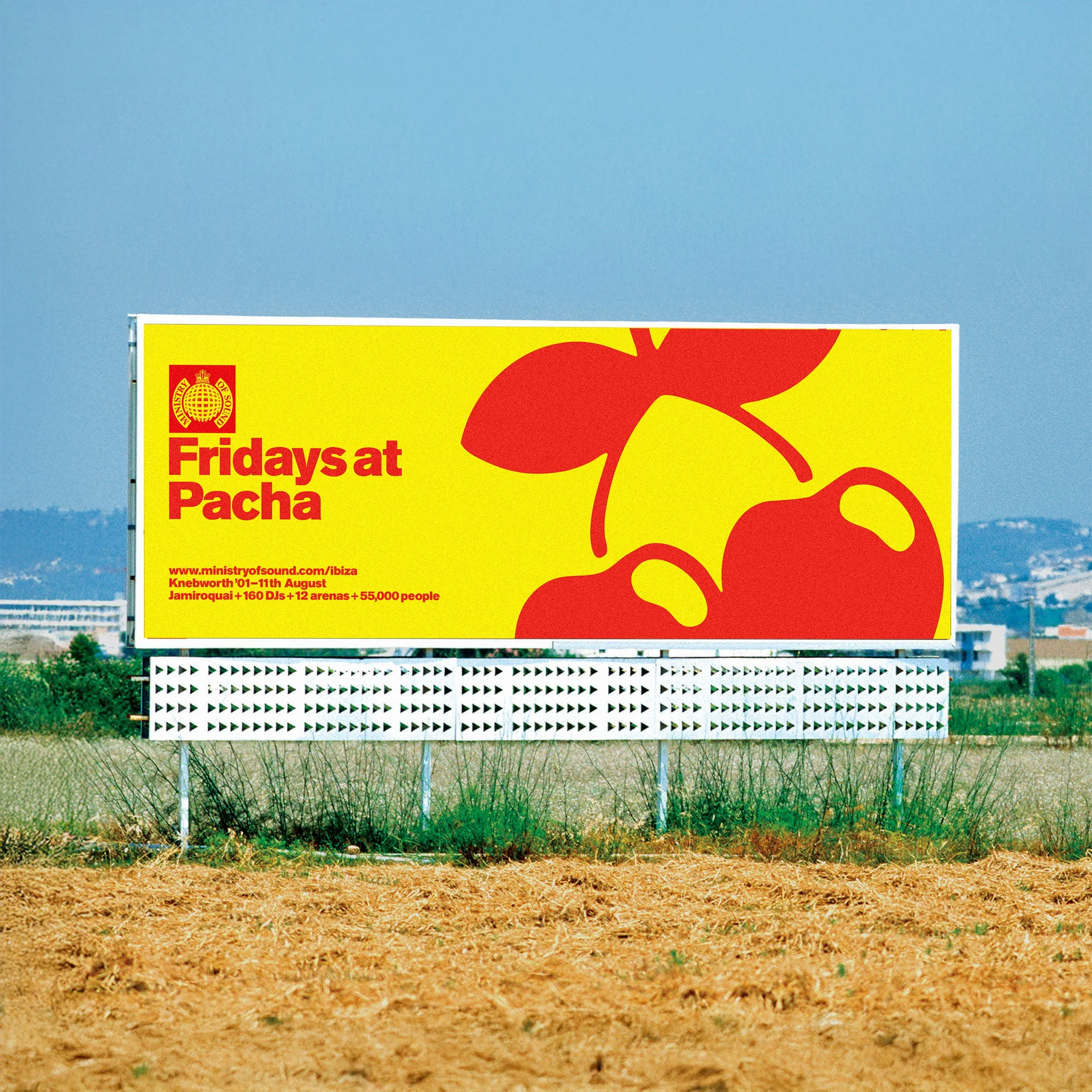 Large outdoor billboard advertising 'Fridays at Pacha' with a yellow background, red cherries and text, located in an open area with some grass and buildings in the distance.