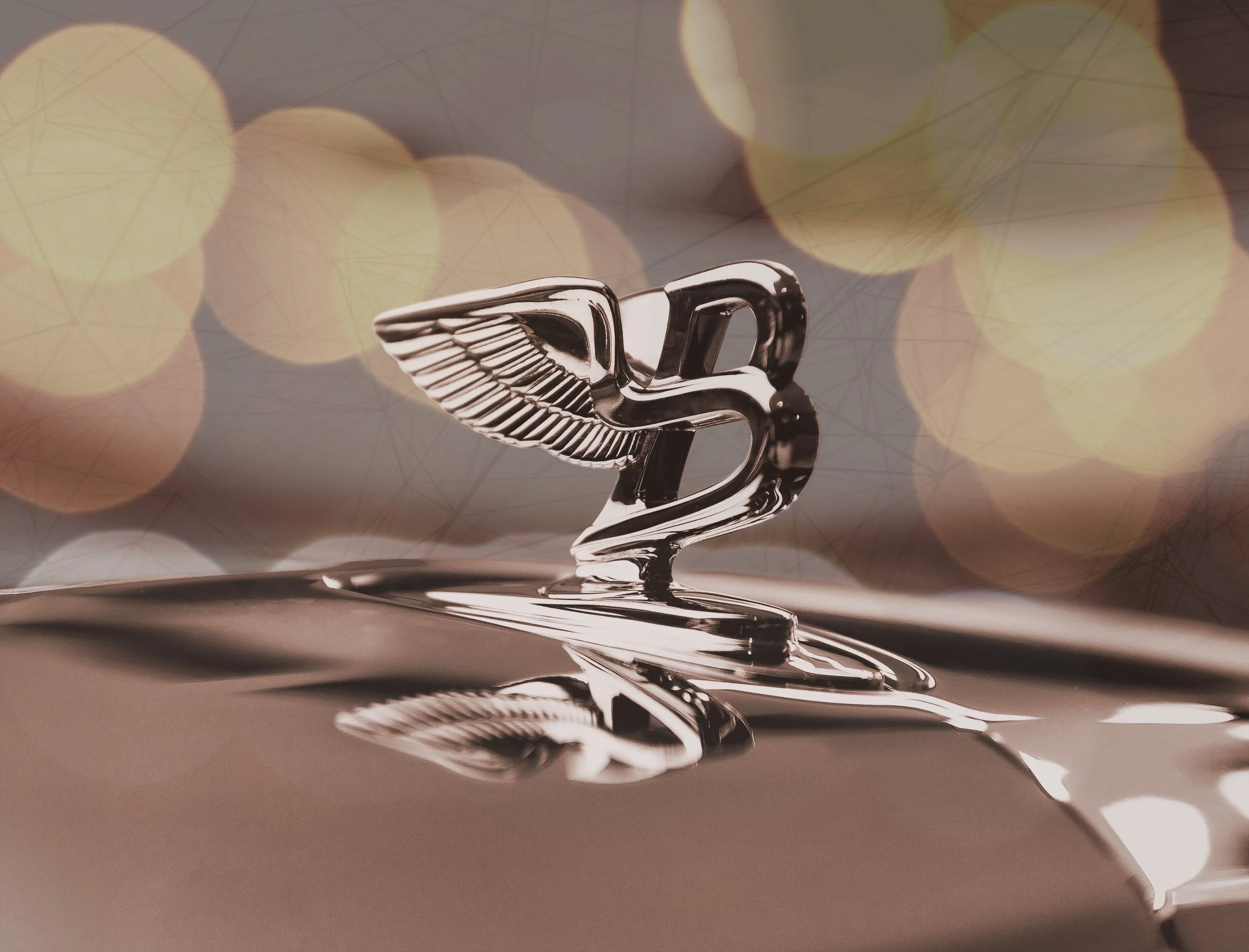 Close-up of a vintage car hood ornament featuring a winged figure, with blurred lights in the background.