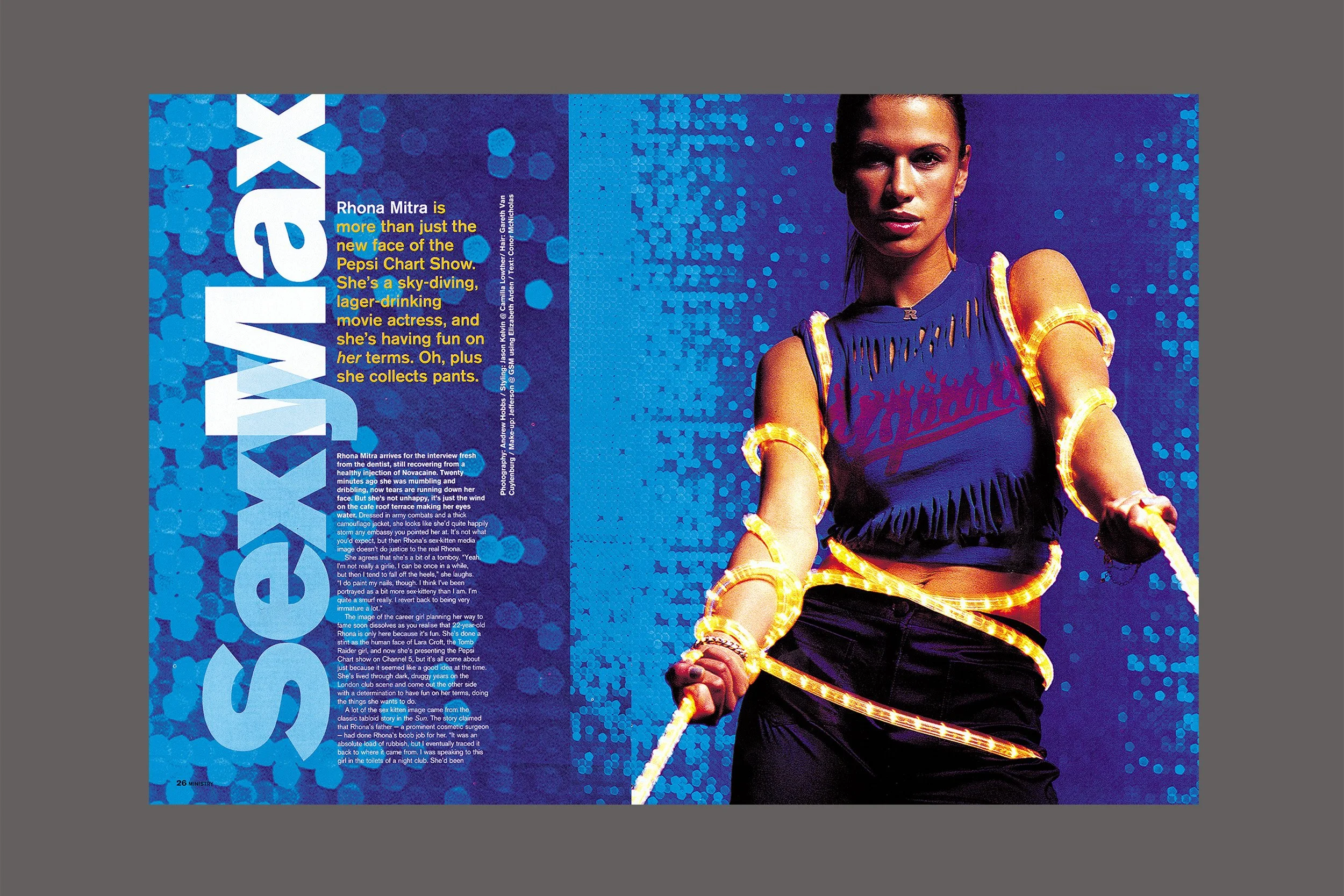 Magazine spread featuring Rhona Mitra, with her name in large blue letters on the left side and a photo of her on the right, dressed in a dark blue outfit with glowing orange accessories against a blue background with a dotted pattern.