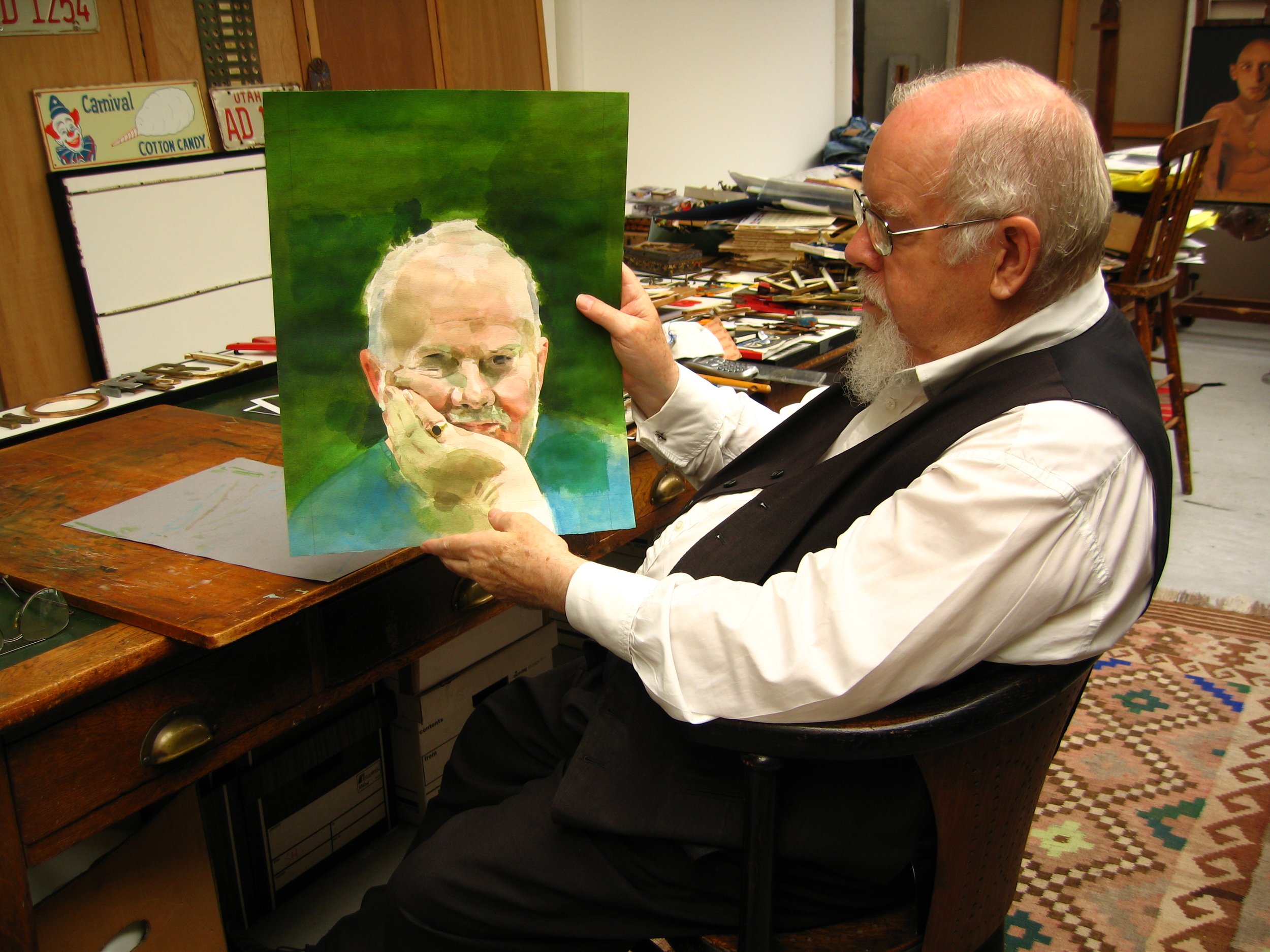 An elderly man with glasses and a white beard, wearing a white shirt and black vest, sits in a wooden chair and holds a portrait painting of a woman with glasses, with a background of green hues. The room has a cluttered wooden work table with art supplies, a colorful patterned rug, and artwork in the background.