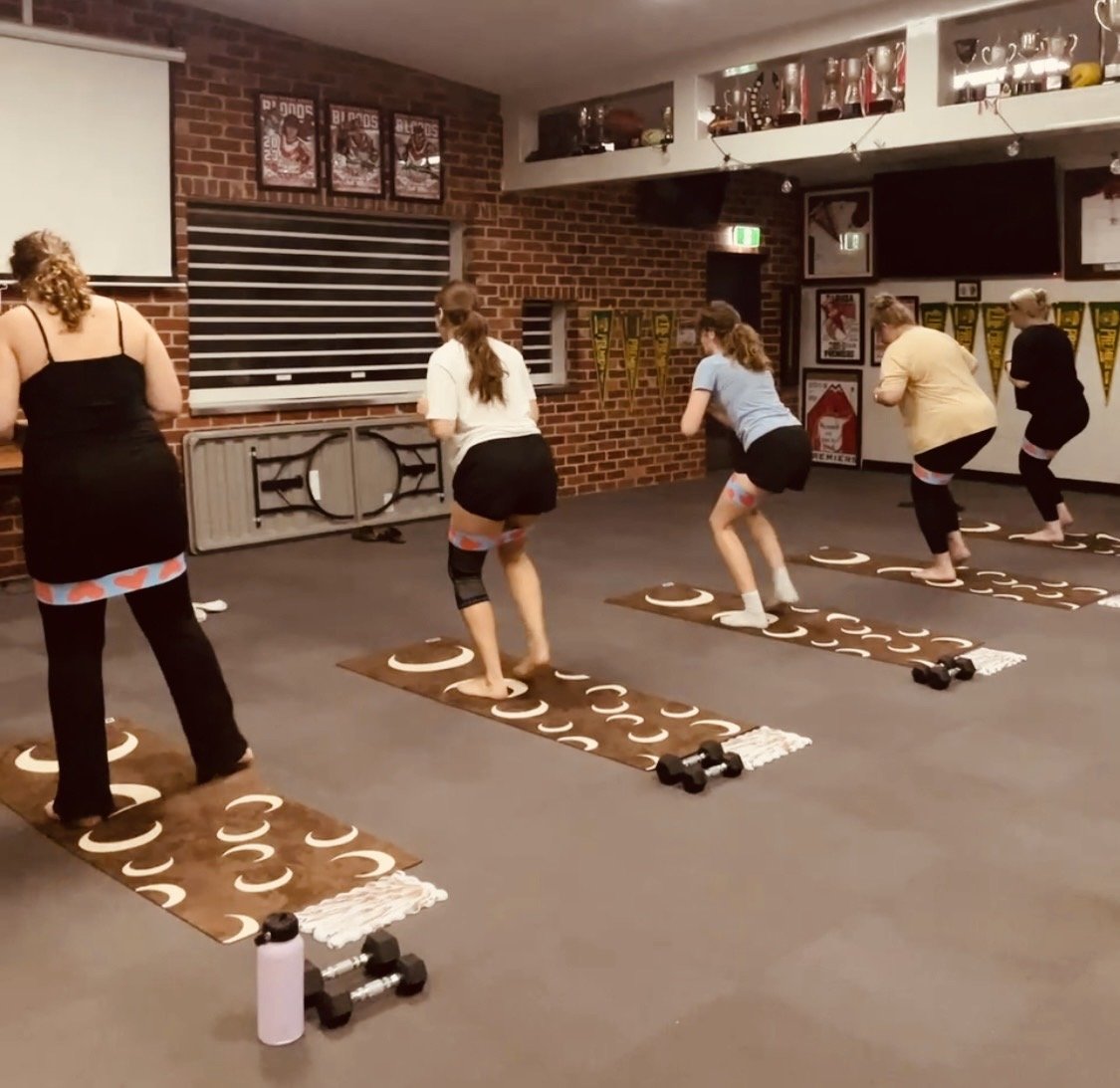 Five women participating in a group mat Pilates class in a football, netball club, performing exercises on Pilates mats with small dumbbells and water bottles nearby.