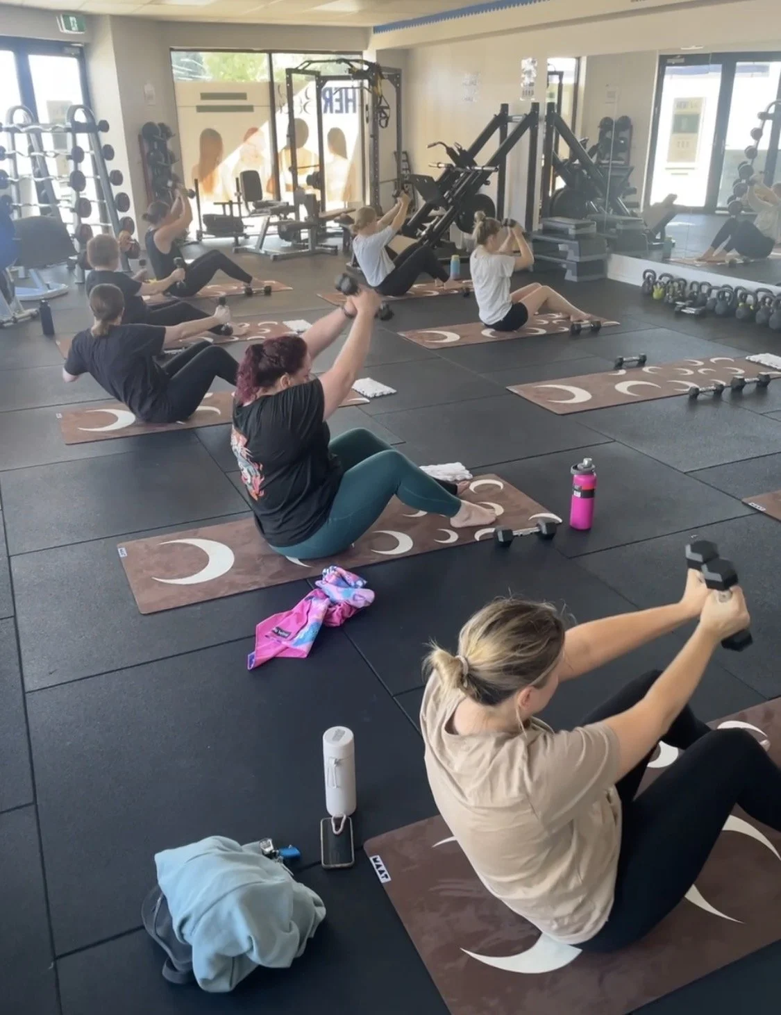 Women participating in a group strength Pilates class in a gym, seated on mats, lifting weights and stretching.