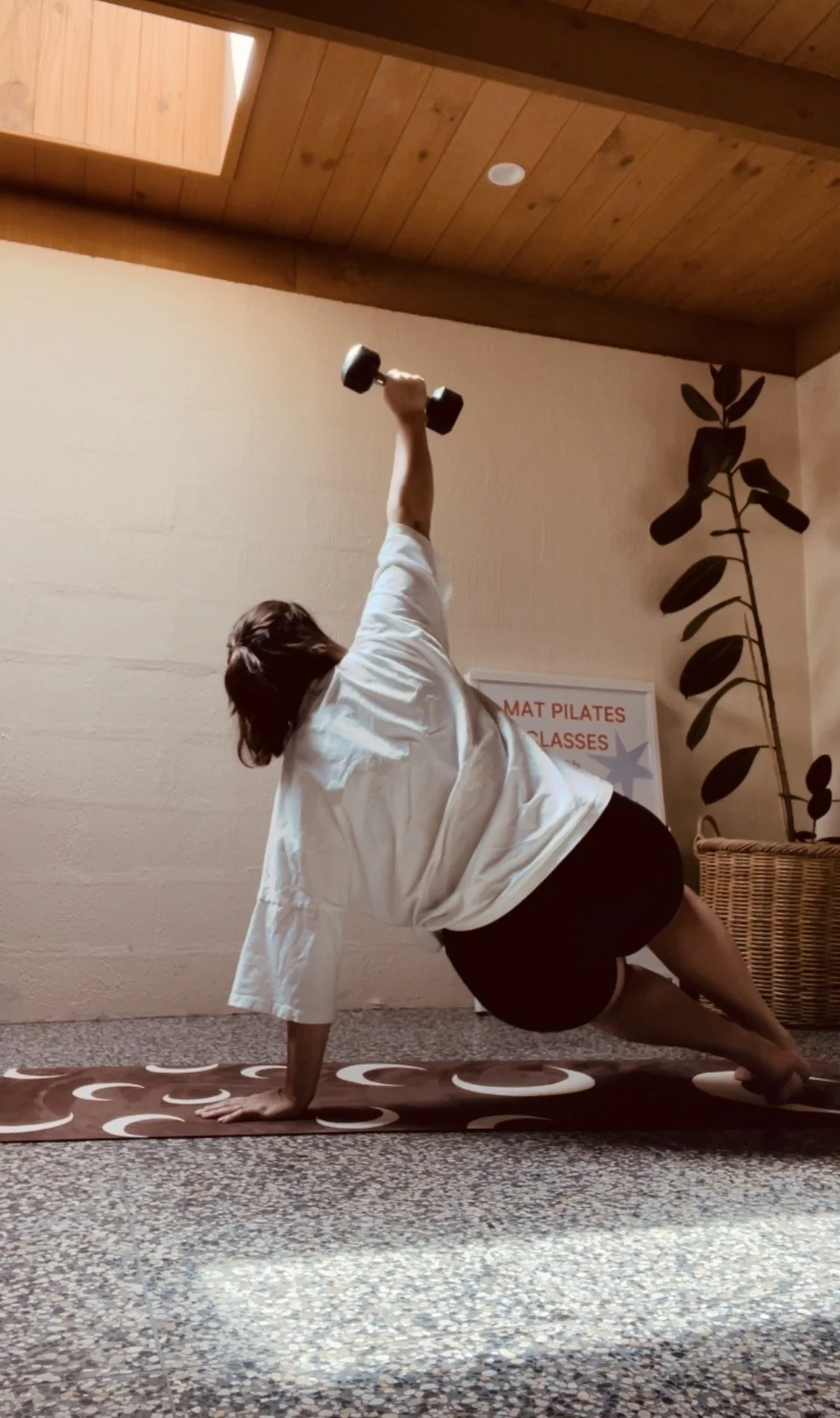 Person practicing mat Pilates on a patterned mat inside a room with wooden ceiling and a plant on the right, holding a dumbbell in one hand while in a side rotation position.