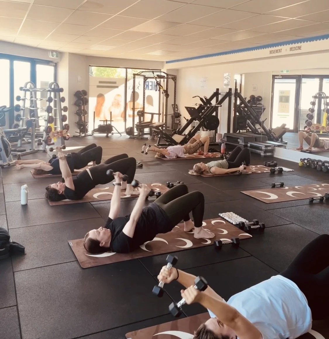 Group of women doing a strength Pilates class with dumbbells on exercise mats in a gym.