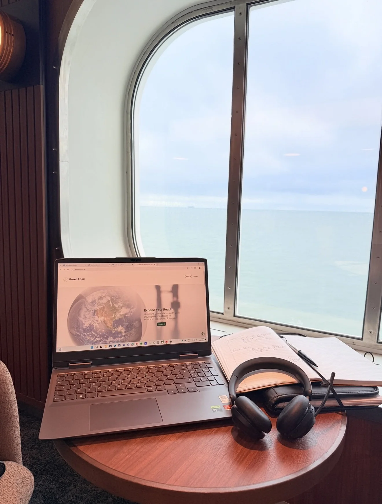 A desk with a laptop displaying a webpage, a notebook, headphones, and a pen. A large window shows a view of the ocean and sky outside.