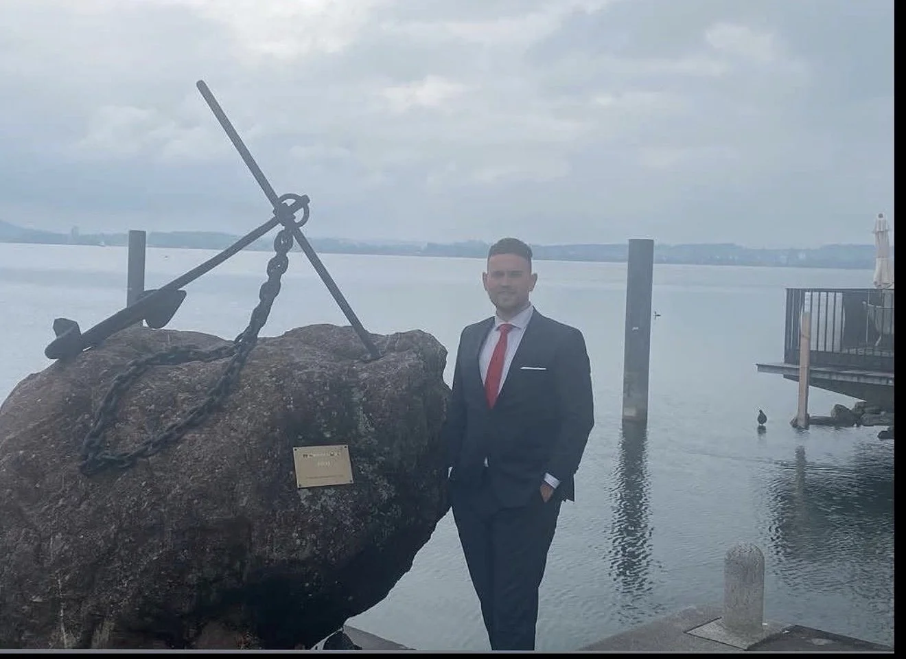 A man standing in a suit with a red tie standing infront of a lake in Switzerland