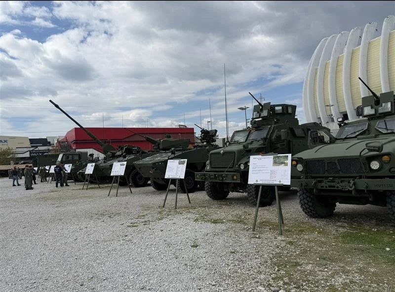 Military vehicles parked outdoors at an exhibition with informational signs in front of each vehicle and people viewing in the background.