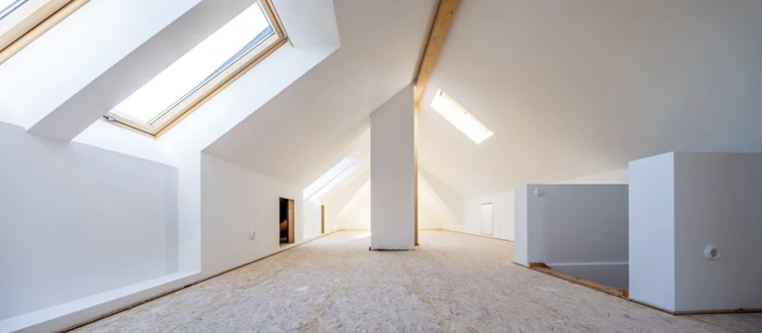 Leeres Dachgeschoss mit weißen Wänden, mehreren Dachfenstern und Holzbalken, noch in Bauphase.