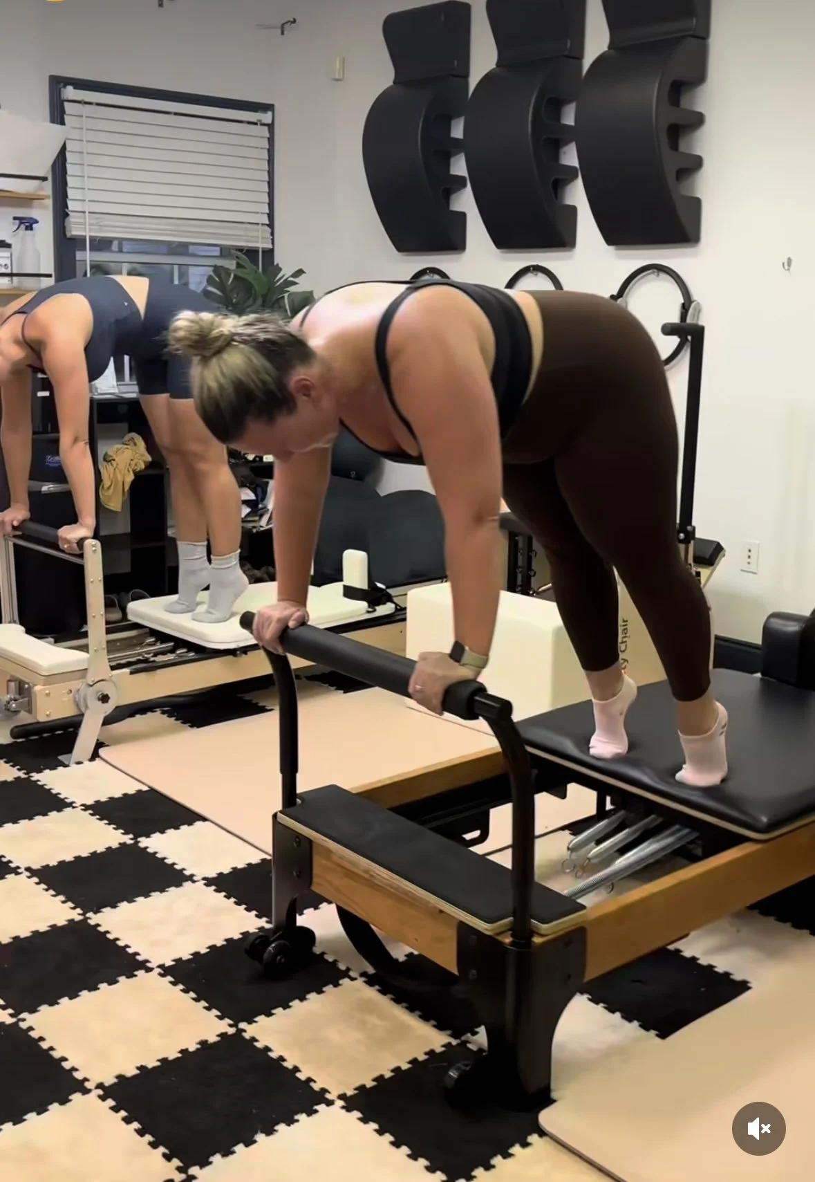 Two women exercising at a Pilates studio; one is doing a plank on a reformer machine, the other is bending forward on a different Pilates equipment.