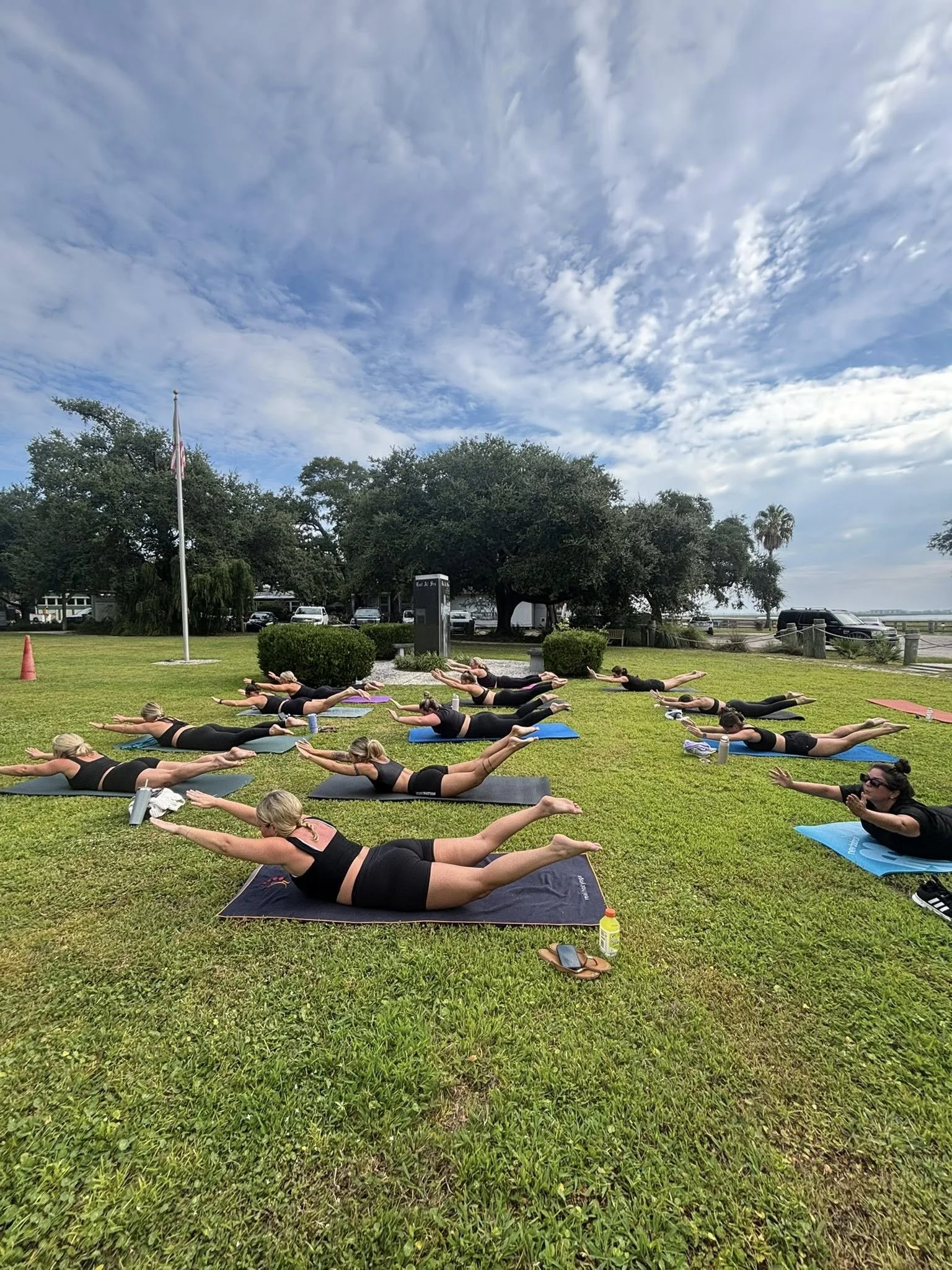 Group of women practicing yoga outdoors on a grassy field under a partly cloudy sky, lying on yoga mats in a plank position with arms extended forward.