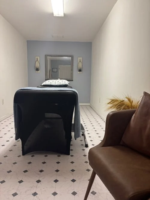 A room with a massage or examination table in the center, a mirror on the back wall, and a brown leather chair on the right side. There are wall-mounted lights and a plant with yellowish leaves in the corner.