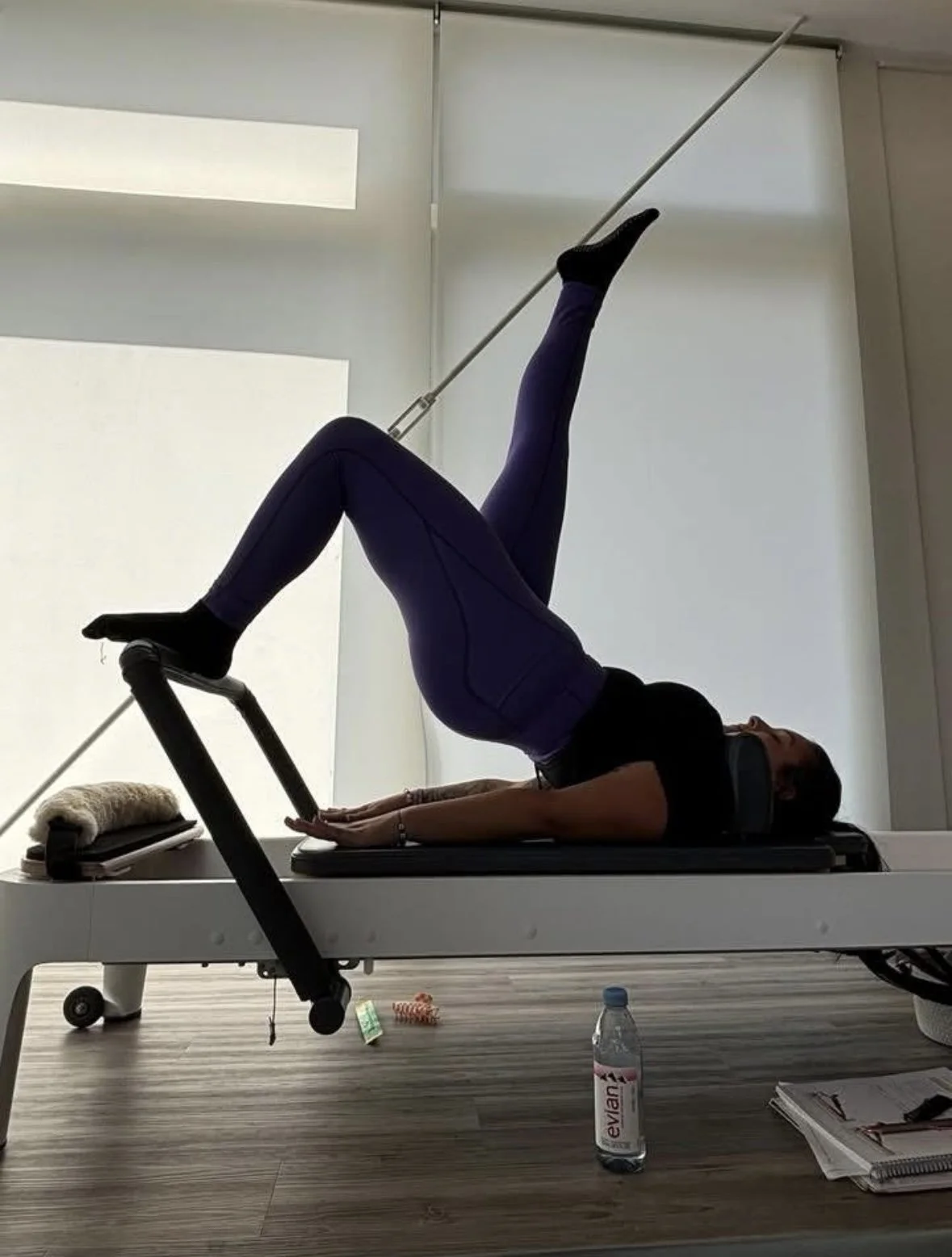 A person performing a yoga pose on a medical examination table. The individual is lying on their back with one leg extended straight up and the other bent, with hands on the table for support. The room has a minimalistic, clean design with a water bo
