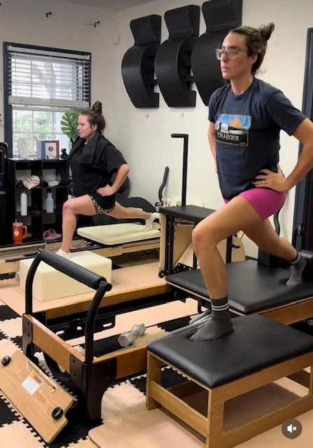 Two women exercising on Pilates reformers in a fitness studio, one in a black outfit and the other in a blue shirt and pink shorts, with a window and fitness equipment in the background.