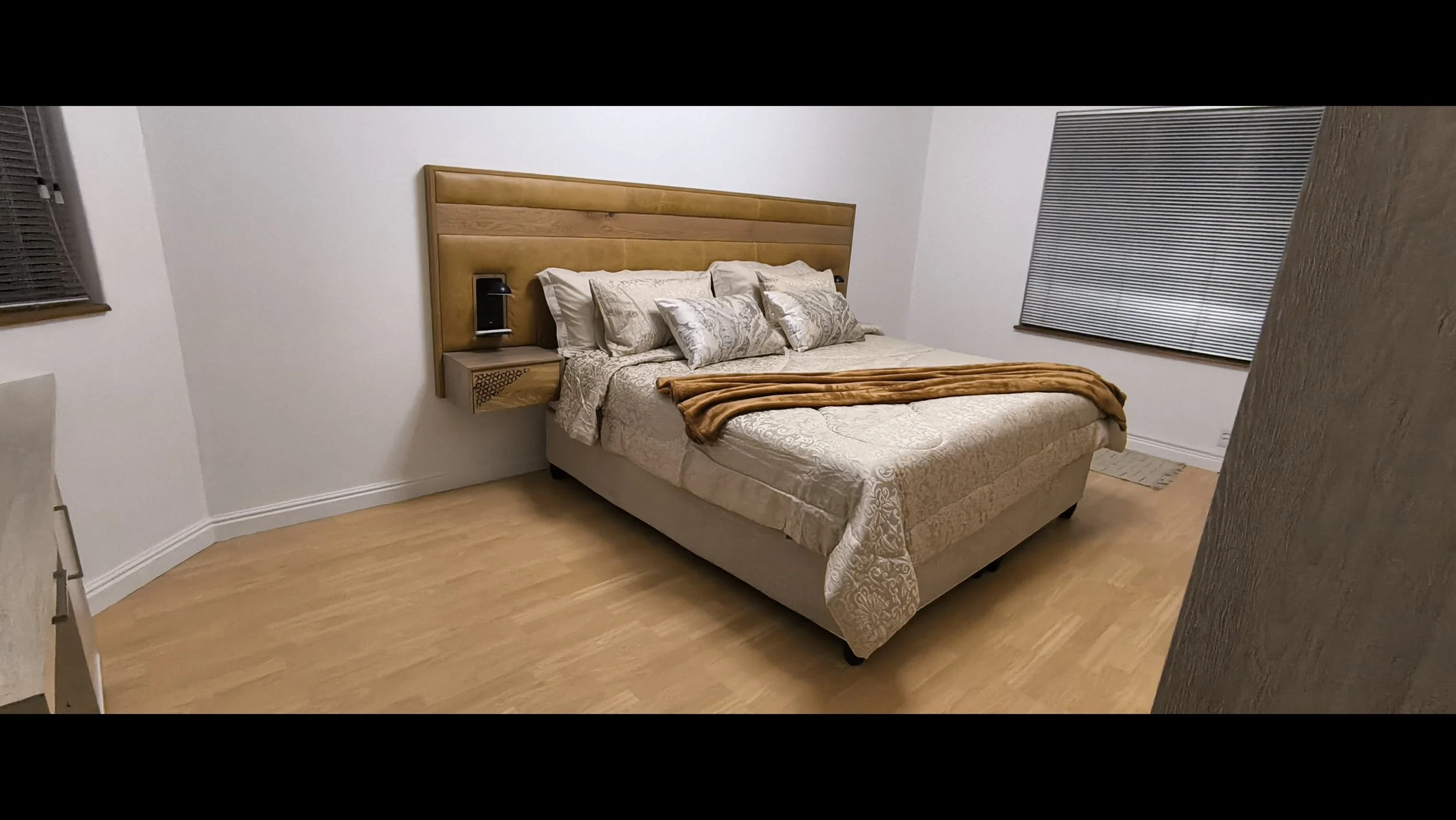 A neatly made bed with multiple decorative pillows, a beige and gold bedspread, and a brown throw blanket. The bed has a wooden headboard with integrated wall-mounted nightlights. There is a window with closed blinds on the right side of the room.