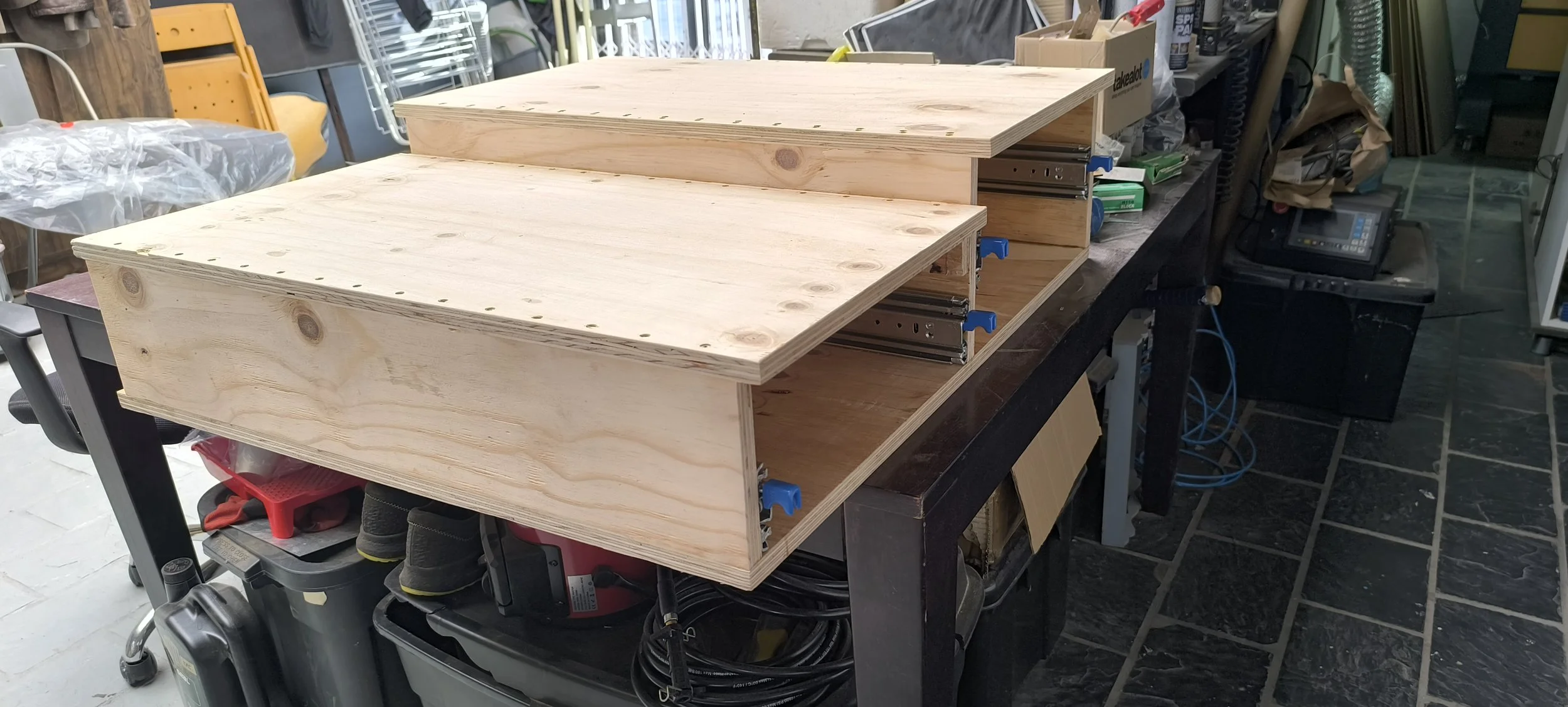Wooden drawer boxes partially assembled on a workshop workbench, with various tools and supplies nearby.
