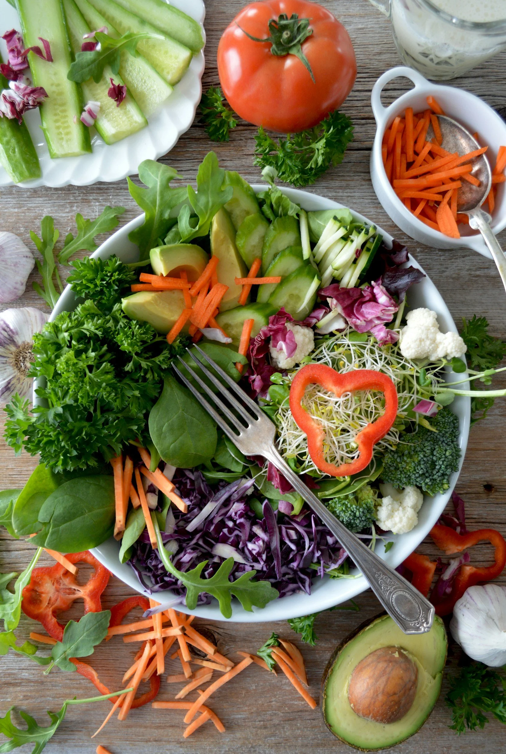 A bowl of fresh salad with cucumber, avocado, cherry tomatoes, carrots, cauliflower, broccoli, red bell pepper, purple cabbage, and various greens on a wooden table, with additional vegetables and a small bowl of sliced carrots nearby.