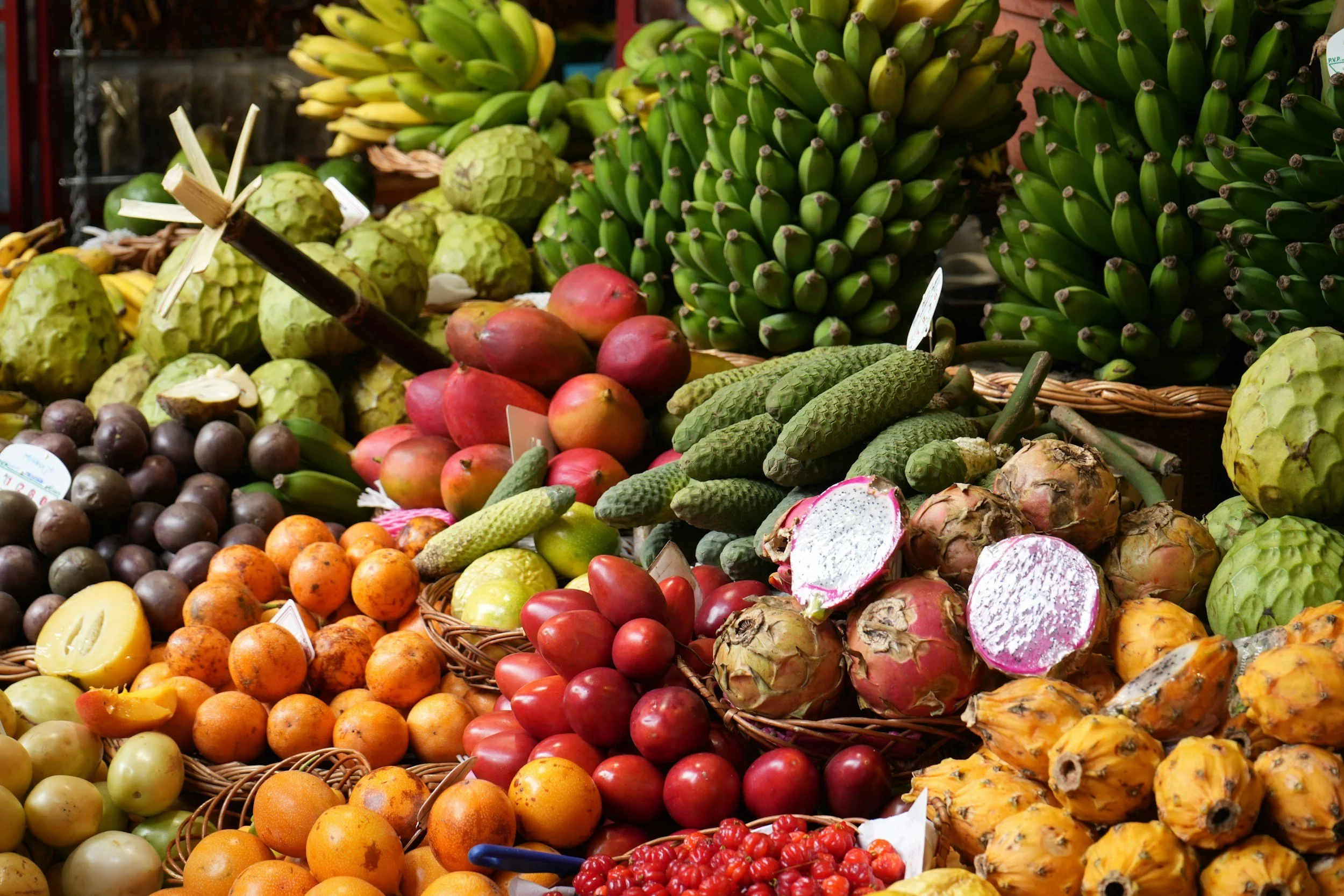 A colorful display of various tropical fruits including bananas, cherimoyas, dragon fruits, papayas, lychees, apples, cucumbers, and tomatoes at a market stall.