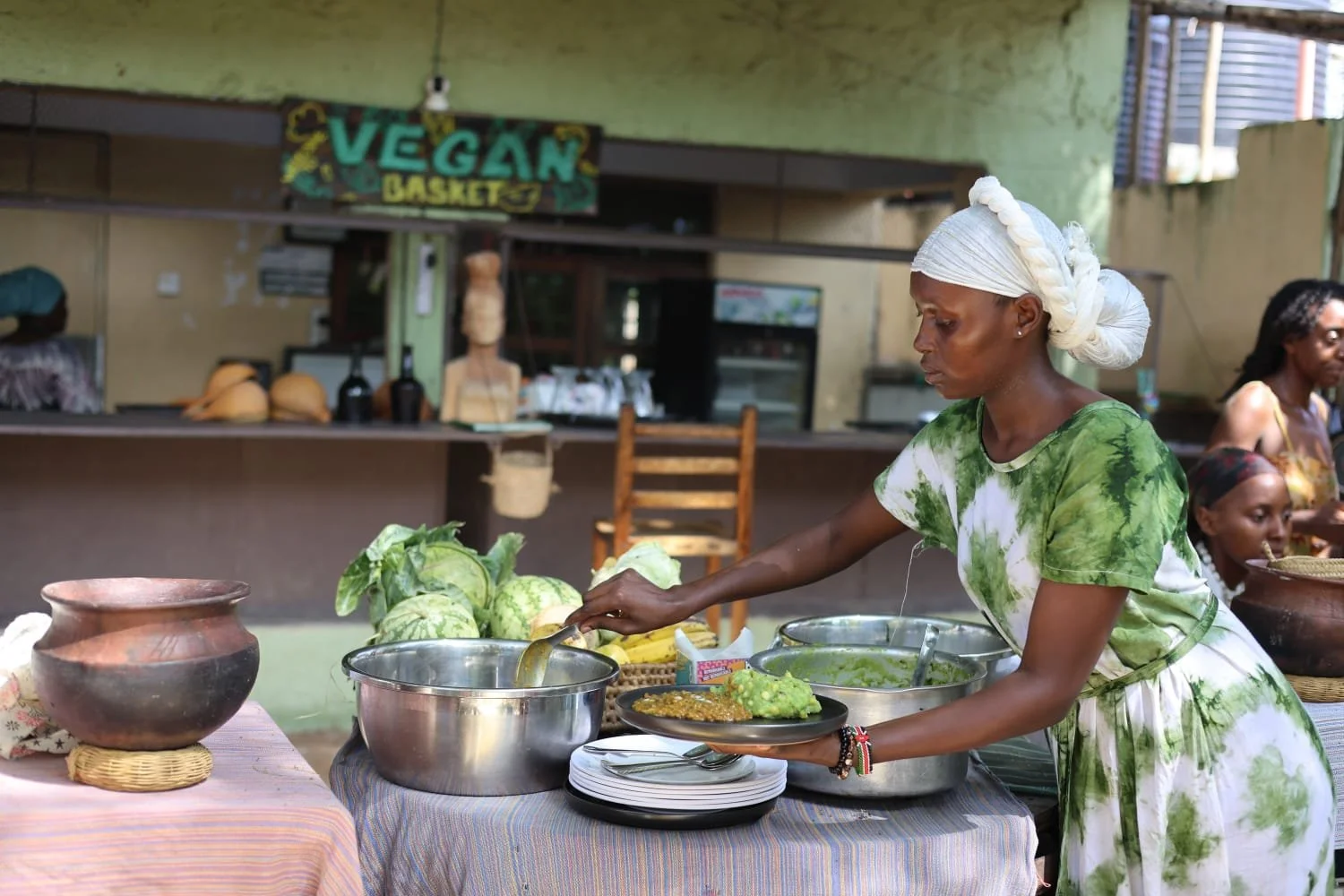 A woman serving food at a table with bowls of vegetables and plates, in front of a sign that reads 'Vegan Basket', in an open-air setting.