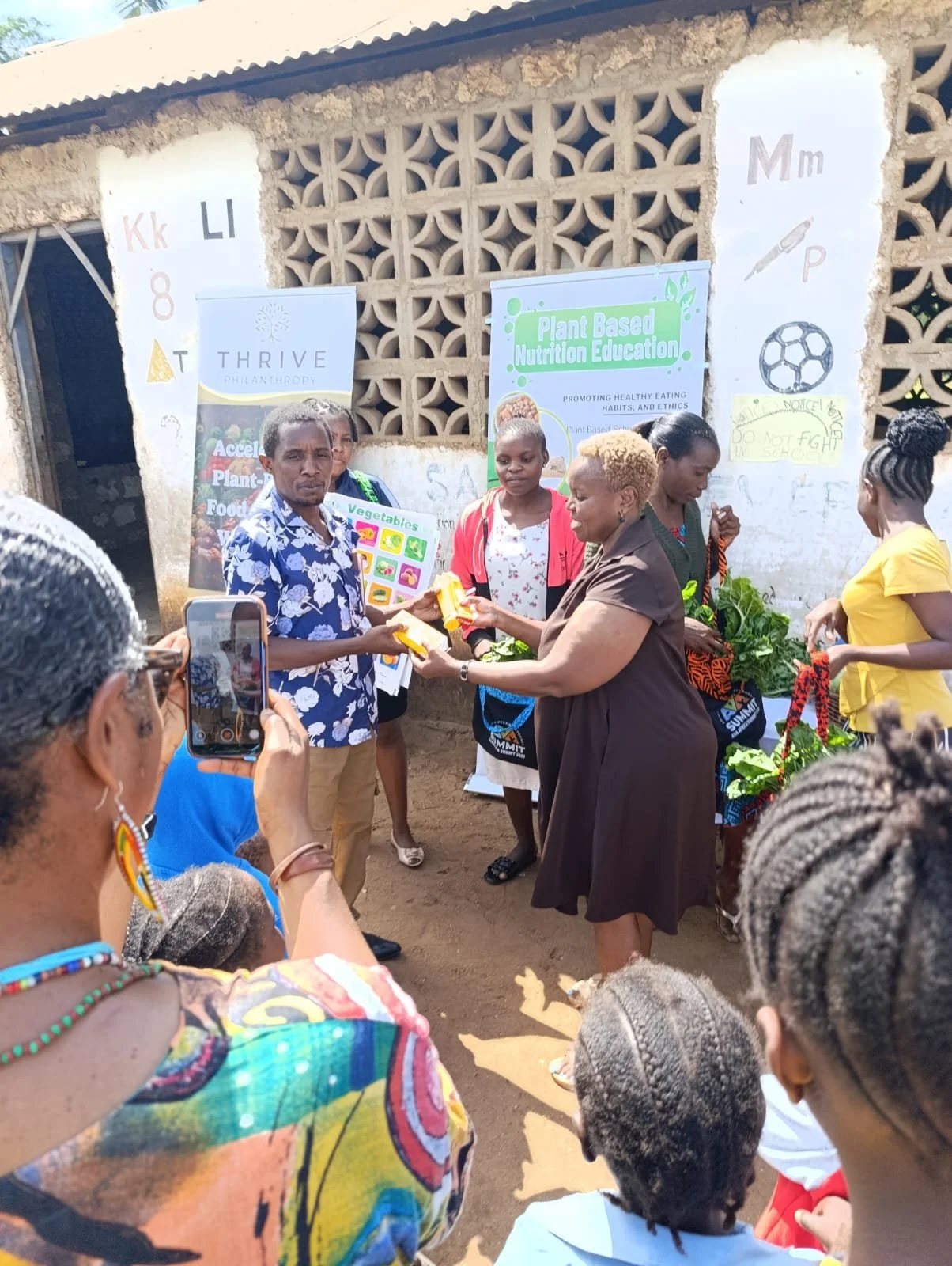 A group of people gathered outdoors during a community event, with a woman handing out items to a man, while others look on. There are signs in the background about plant-based nutrition education, and some children are present in the foreground.